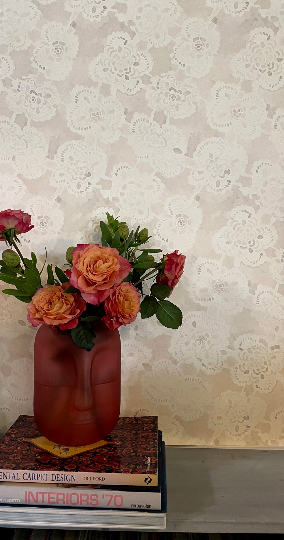 Vase shaped like a face with flowers on top of books against a patterned wall.