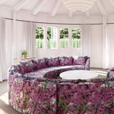 A large round sofa upholstered in a painterly botanical print in purple, green and brown stands in the middle of a sunny room.