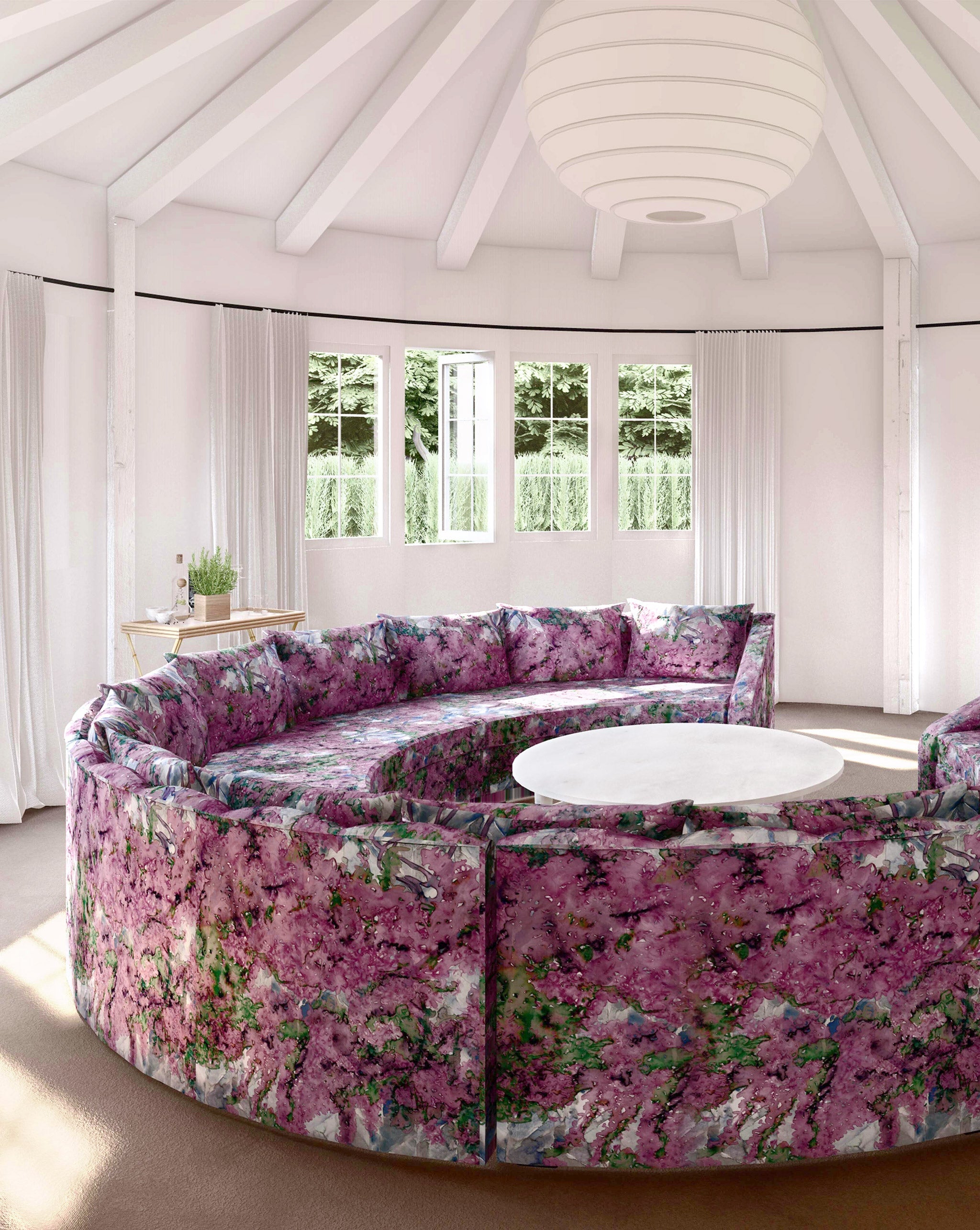 A large round sofa upholstered in a painterly botanical print in purple, green and brown stands in the middle of a sunny room.