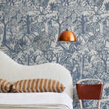 A modernist bed, hanging lamp and chair stand in front of a wall papered in a playful jungle print in blue and cream.