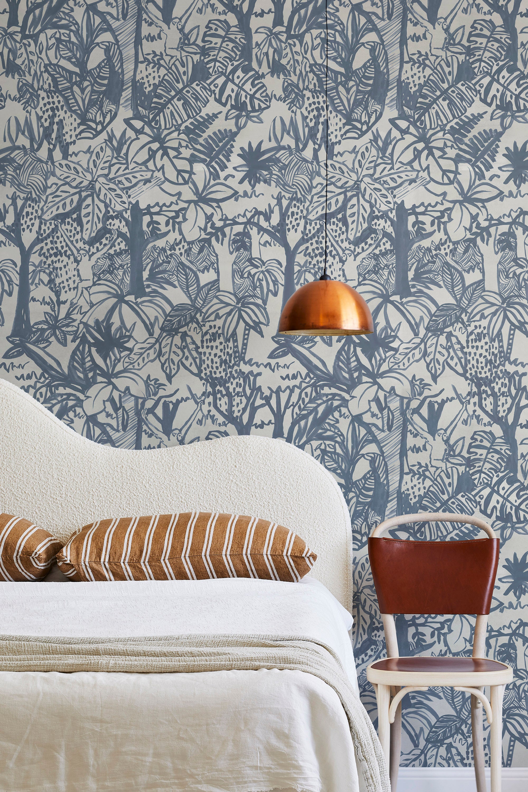 A modernist bed, hanging lamp and chair stand in front of a wall papered in a playful jungle print in blue and cream.