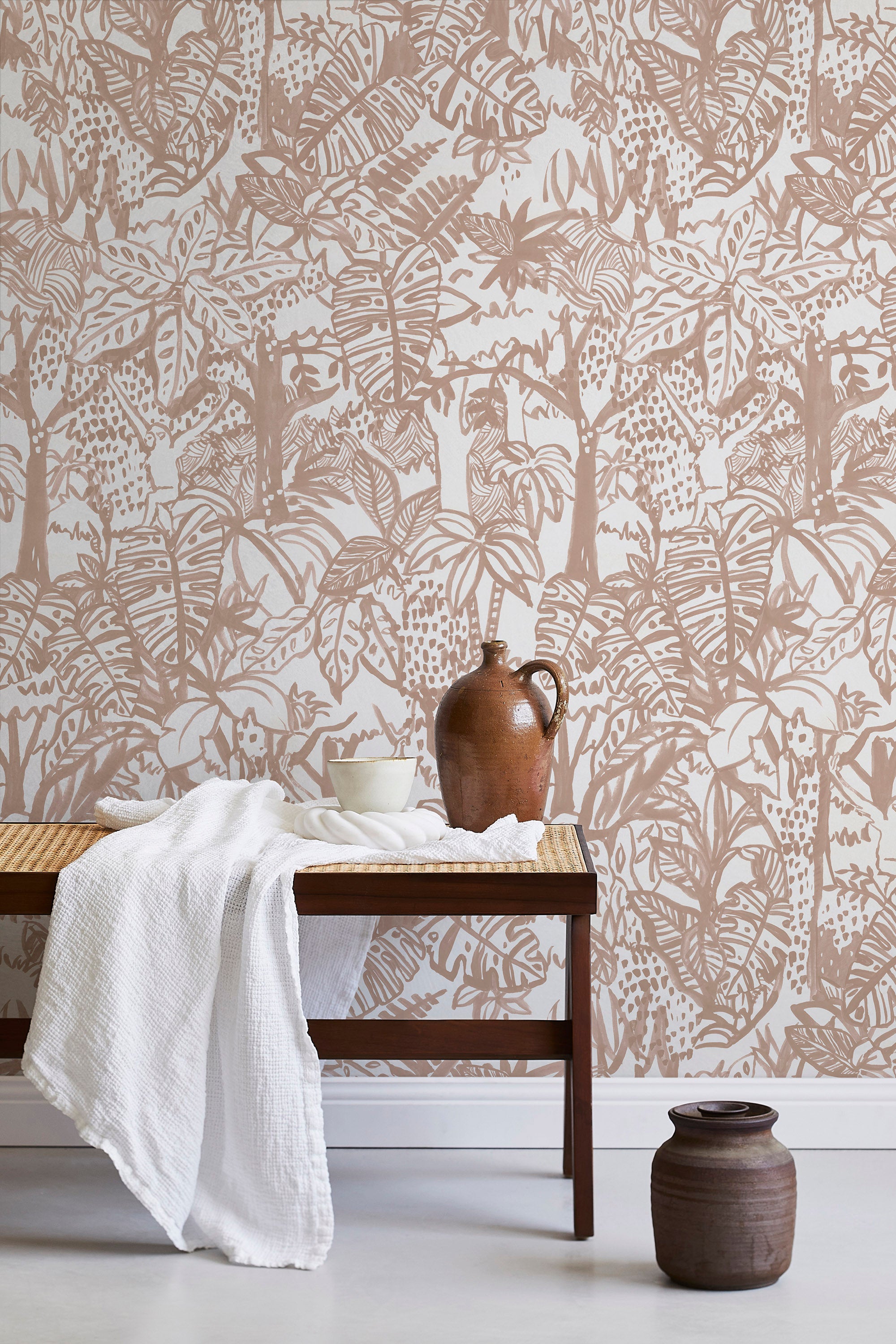 A bench with a towel and clay vases stands in front of a wall papered in a playful jungle print in blush and cream.