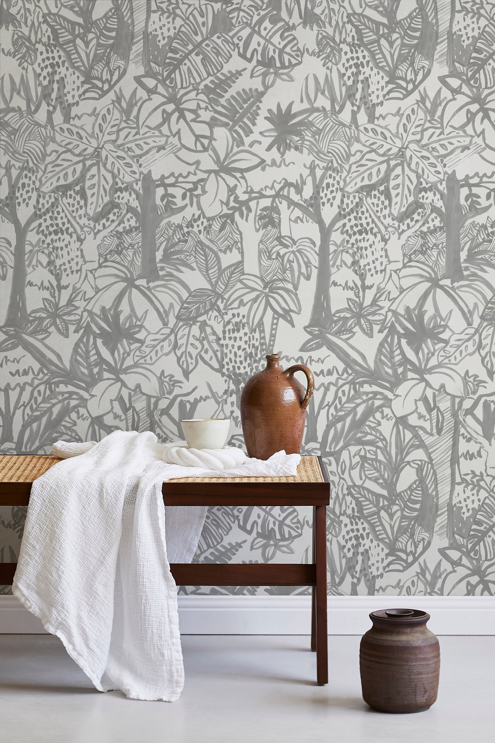 A bench with a towel and clay vases stands in front of a wall papered in a playful jungle print in gray and cream.