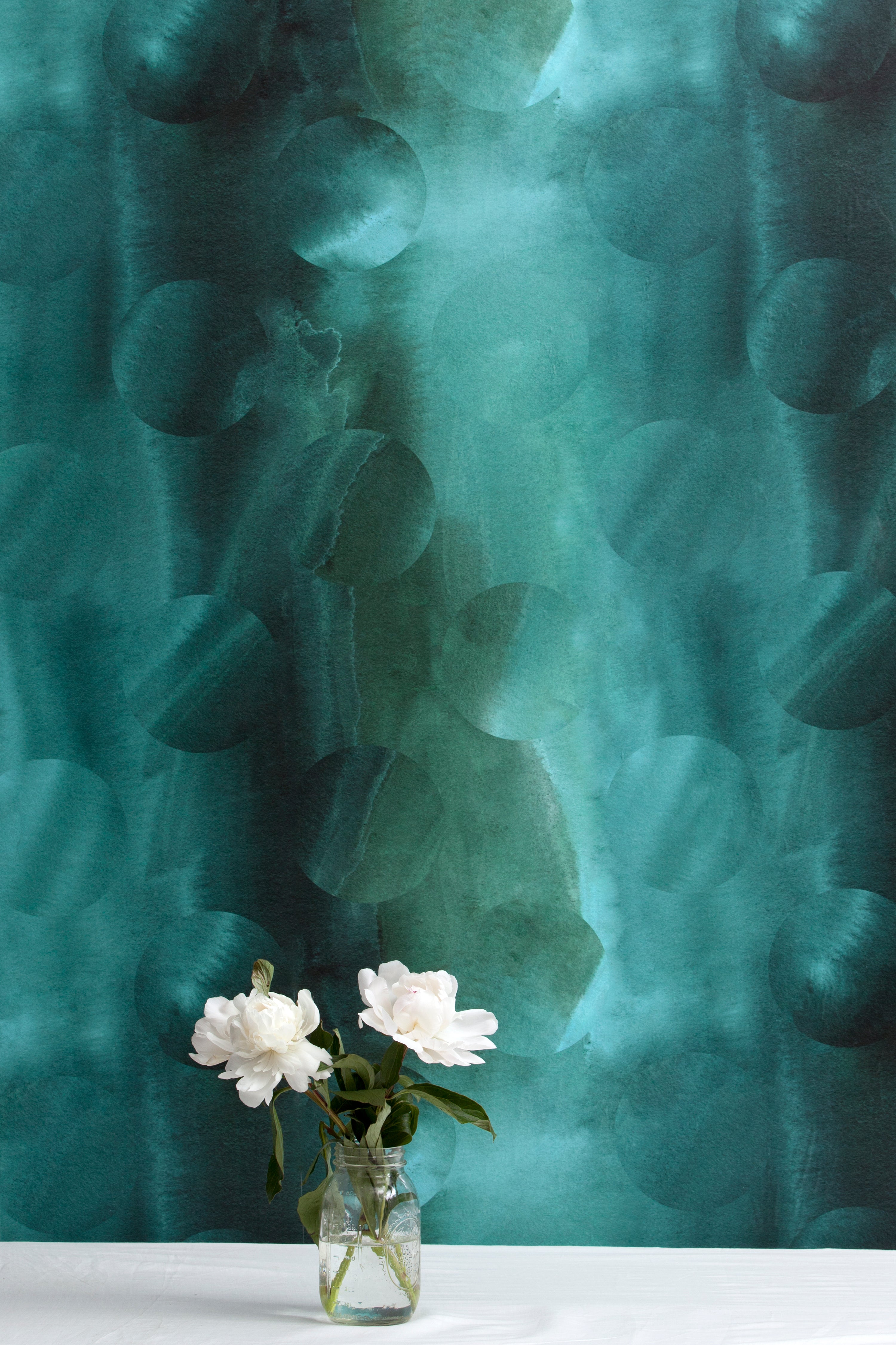 A vase of flowers stands in front of a wall papered in an abstract curvilinear pattern in mottled turquoise and black.