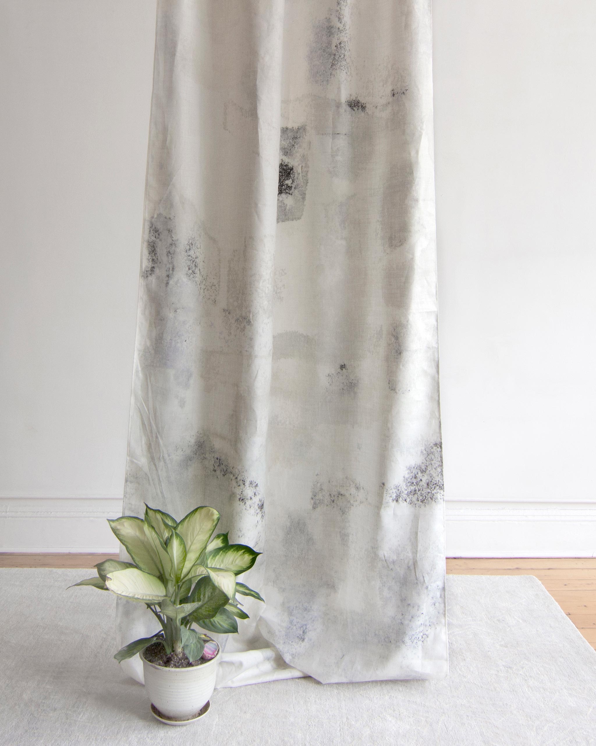 A potted plant sits on the floor in front of a curtain in a minimal splatter print in gray on a cream field.