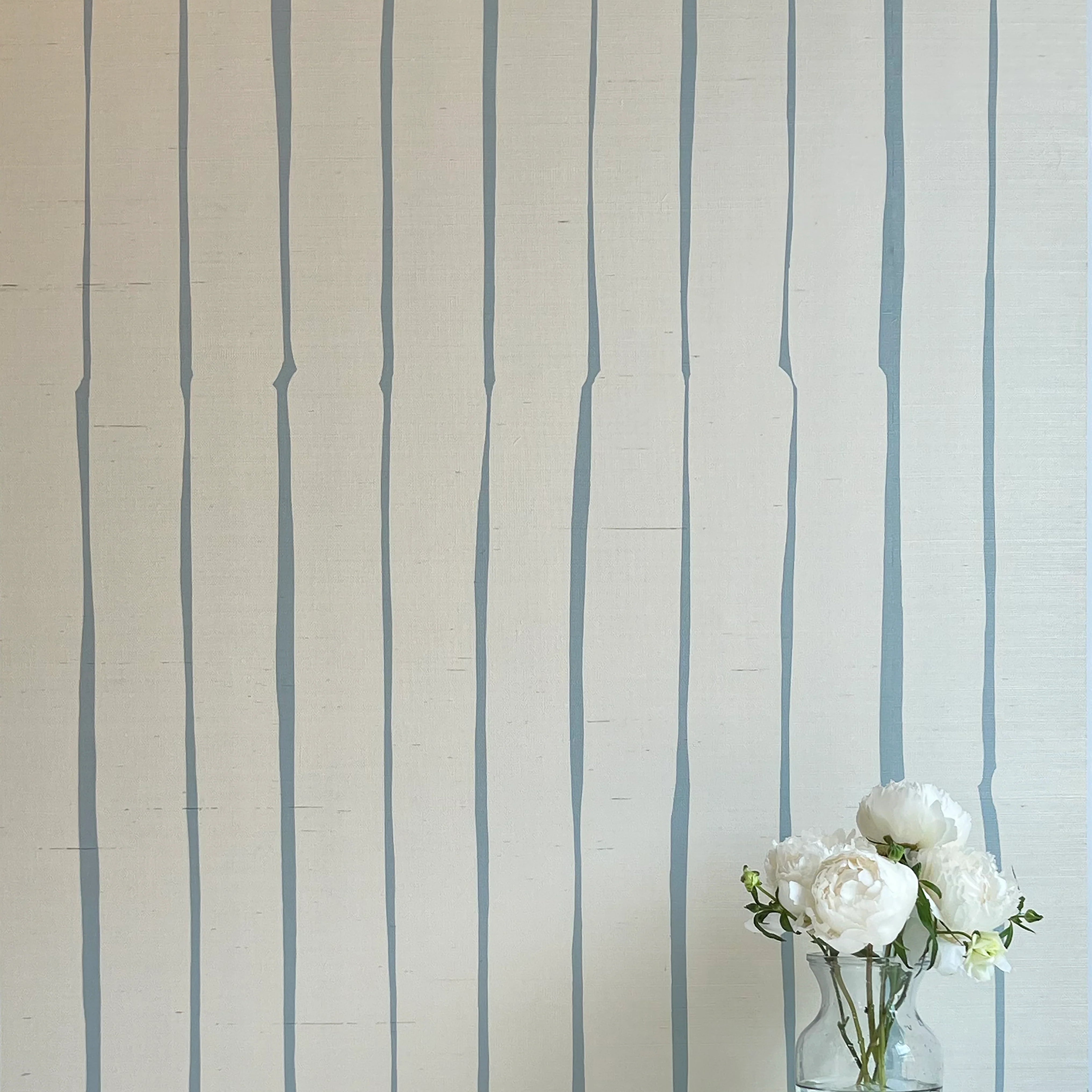 A vase of flowers stands in front of a wall covered in a playful irregular stripe print in blue on a cream field.