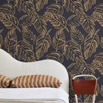 A modernist bed, hanging lamp and chair stand in front of a wall papered in a painterly leaf print in gold and navy.