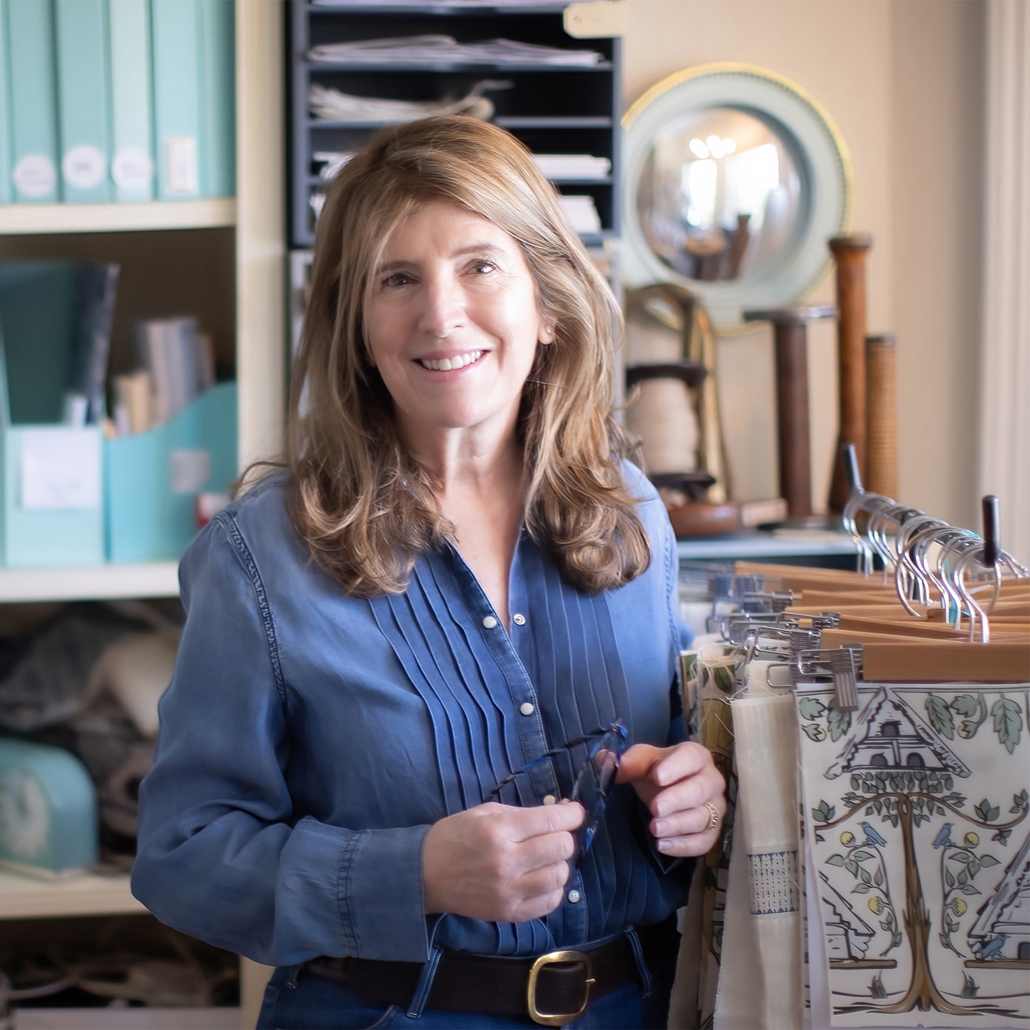 A woman with wavy brown hair and wearing a blue pleated button-down stands next to a rack with swatches of interior fabrics.