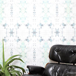 A chair and plant stand in front of a wall papered in an abstract ink blot print in gray and green on a cream field.