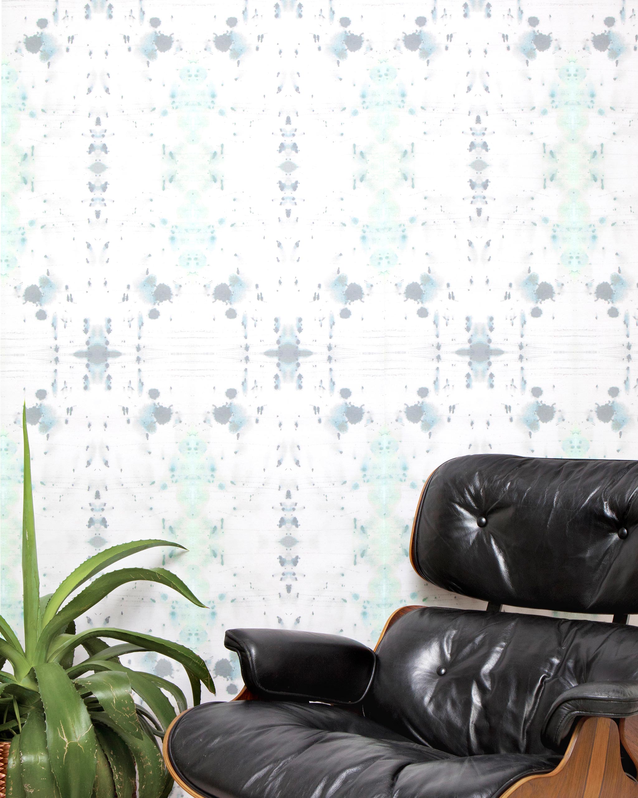 A chair and plant stand in front of a wall papered in an abstract ink blot print in gray and green on a cream field.