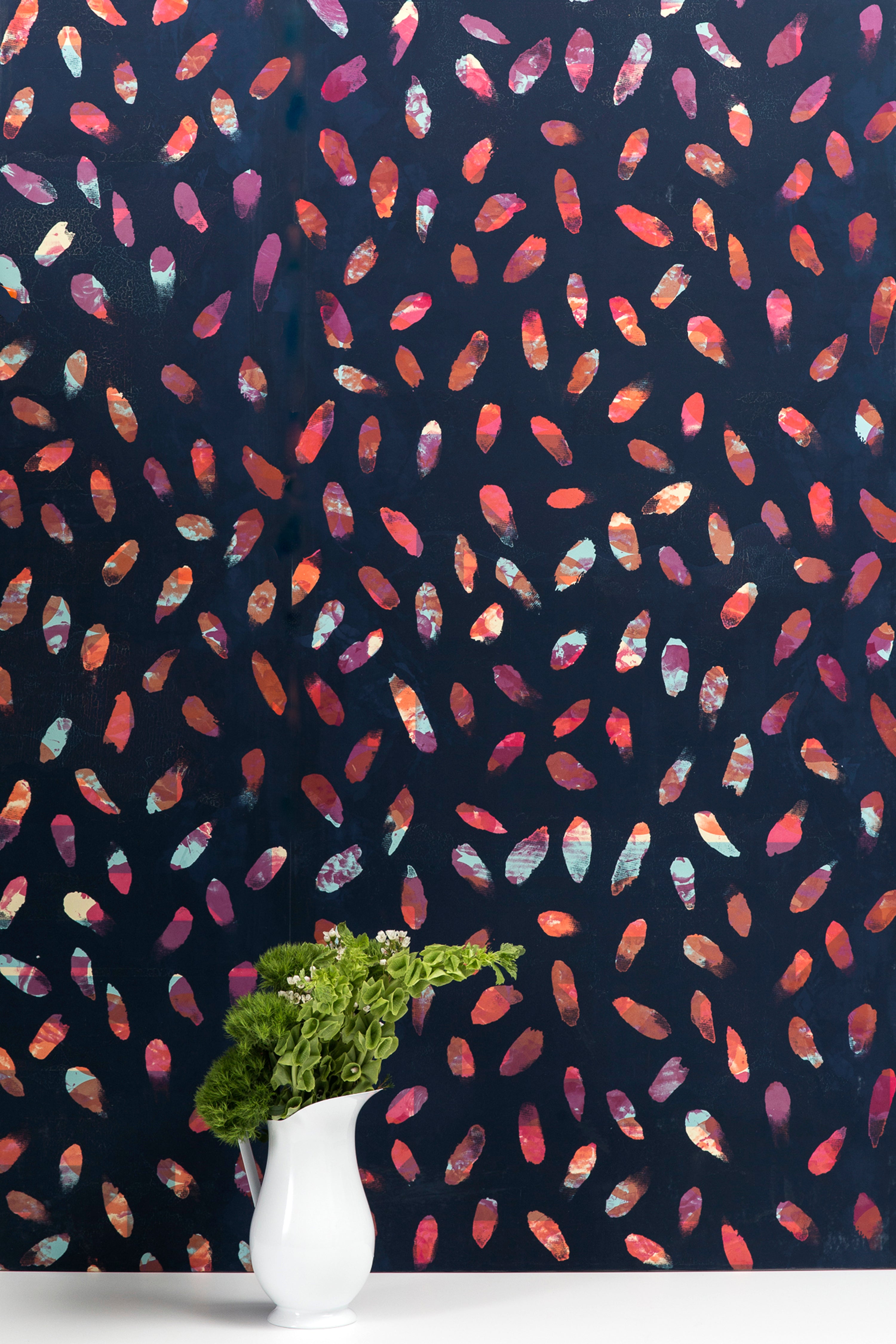 A vase of flowers stands in front of a wall covered in a playful paint blotch print in blue, pink and orange on a navy field.