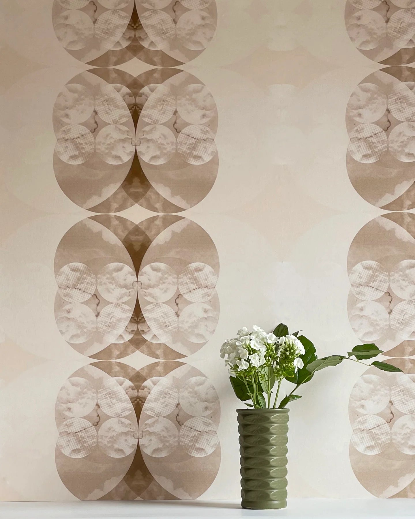 A vase of flowers stands in front of a wall covered in a cloudy ink blot print in shades of brown on a cream field.