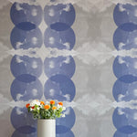 A vase of flowers stands in front of a wall covered in a cloudy ink blot print in shades of blue and gray on a cream field.