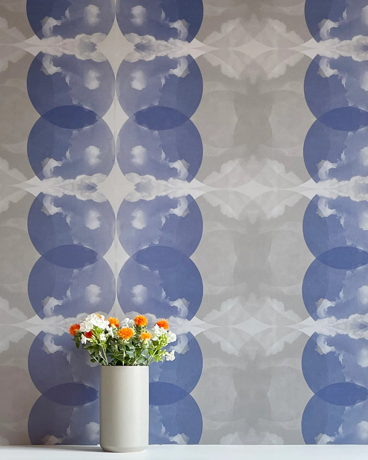 A vase of flowers stands in front of a wall covered in a cloudy ink blot print in shades of blue and gray on a cream field.