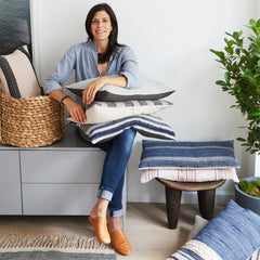 A brown-haired woman in jeans and a button down sits on a bench with a pile of throw pillows on her lap.