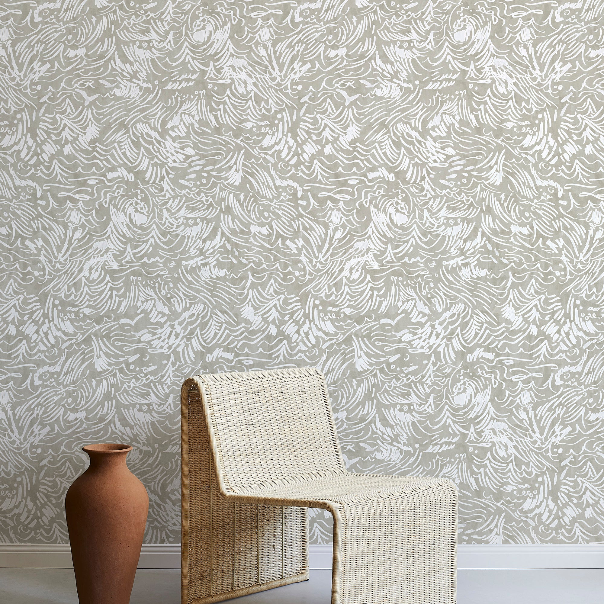 A straw chair and clay pot stand in front of a wall papered in a painterly wave print in white and tan.