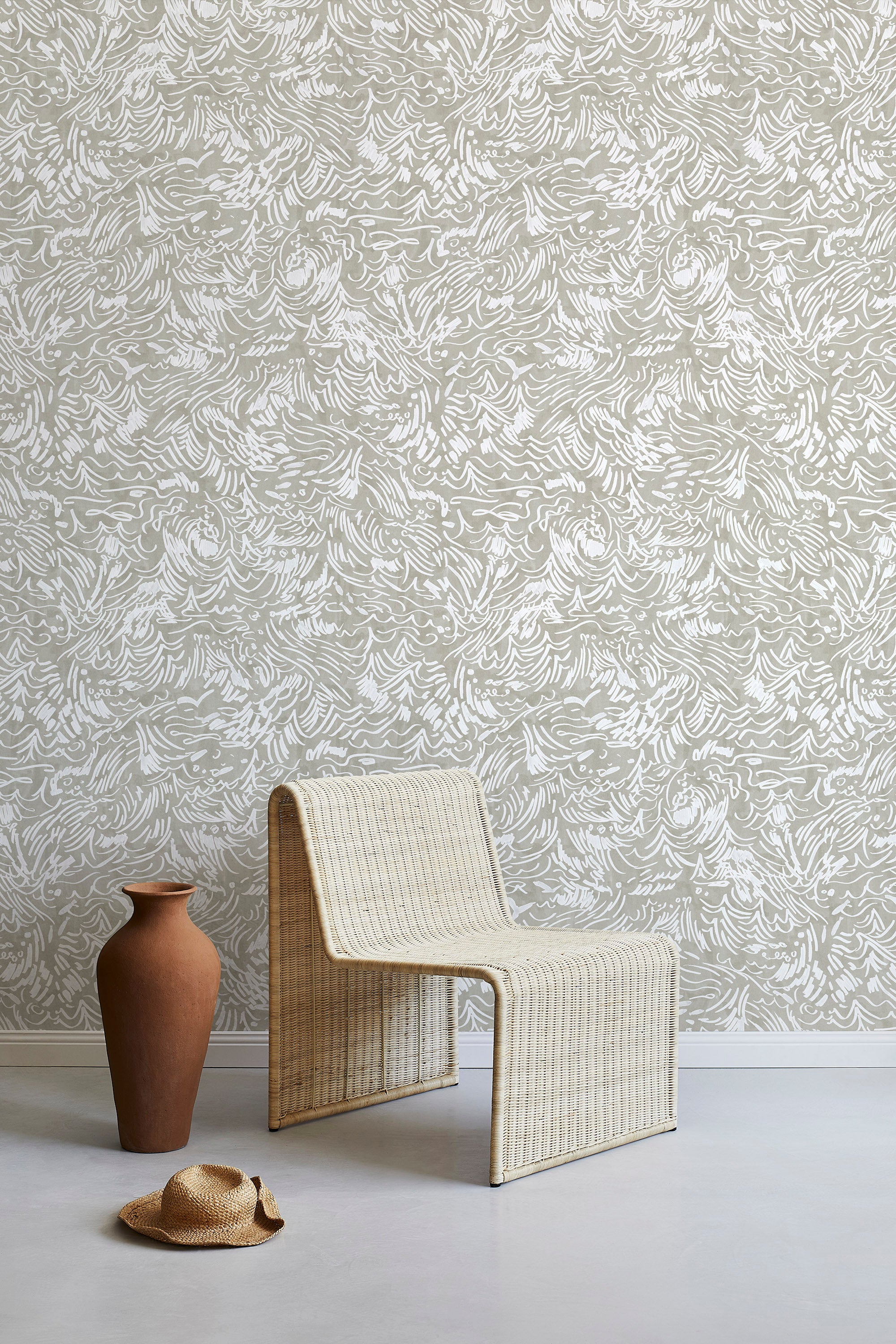 A straw chair and clay pot stand in front of a wall papered in a painterly wave print in white and tan.