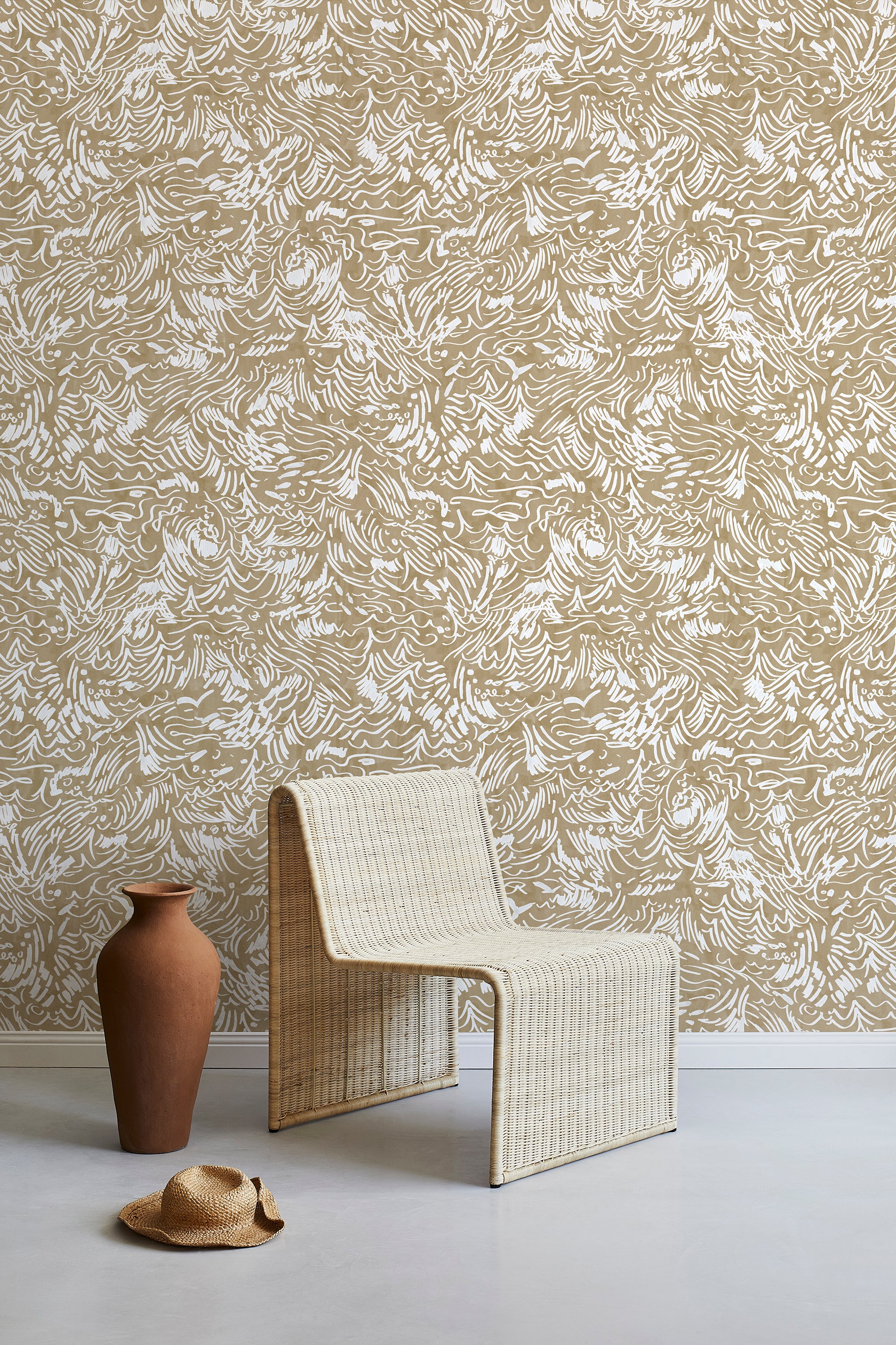 A straw chair and clay pot stand in front of a wall papered in a painterly wave print in white and light brown.