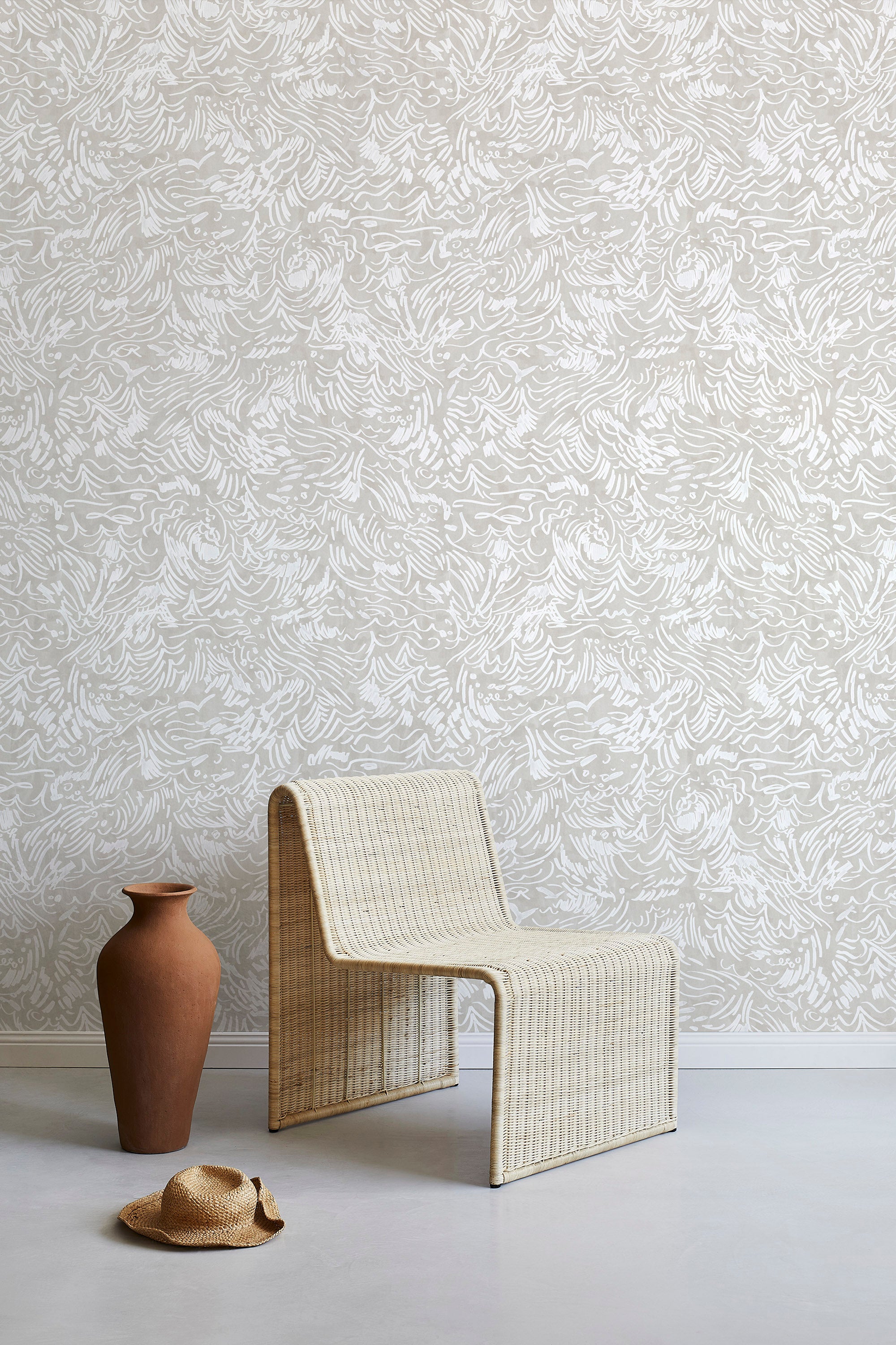 A straw chair and clay pot stand in front of a wall papered in a painterly wave print in white and cream.