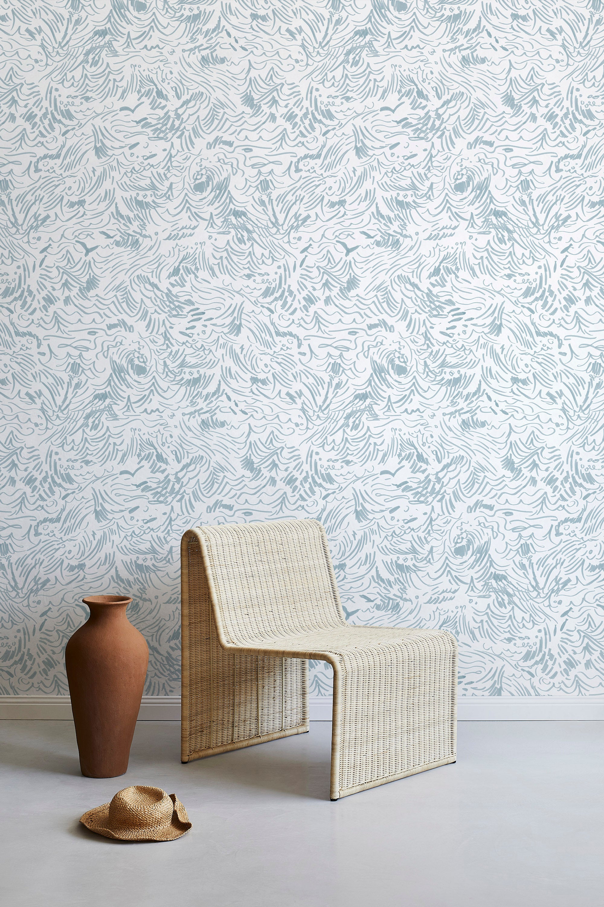 A straw chair and clay pot stand in front of a wall papered in a painterly wave print in light blue and white.