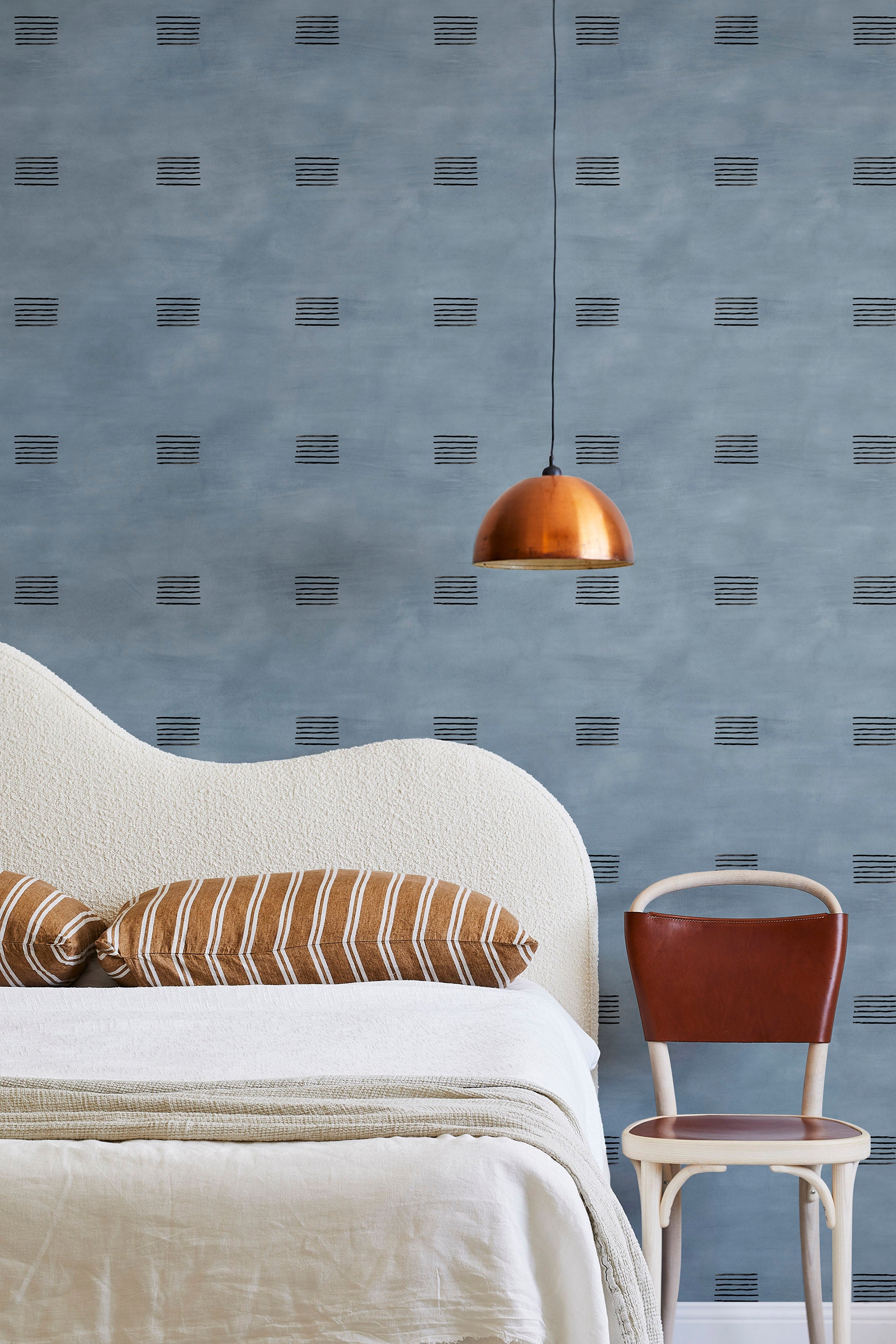 A modernist bed, hanging lamp and chair stand in front of a wall papered in a gridded dash print in black and blue.