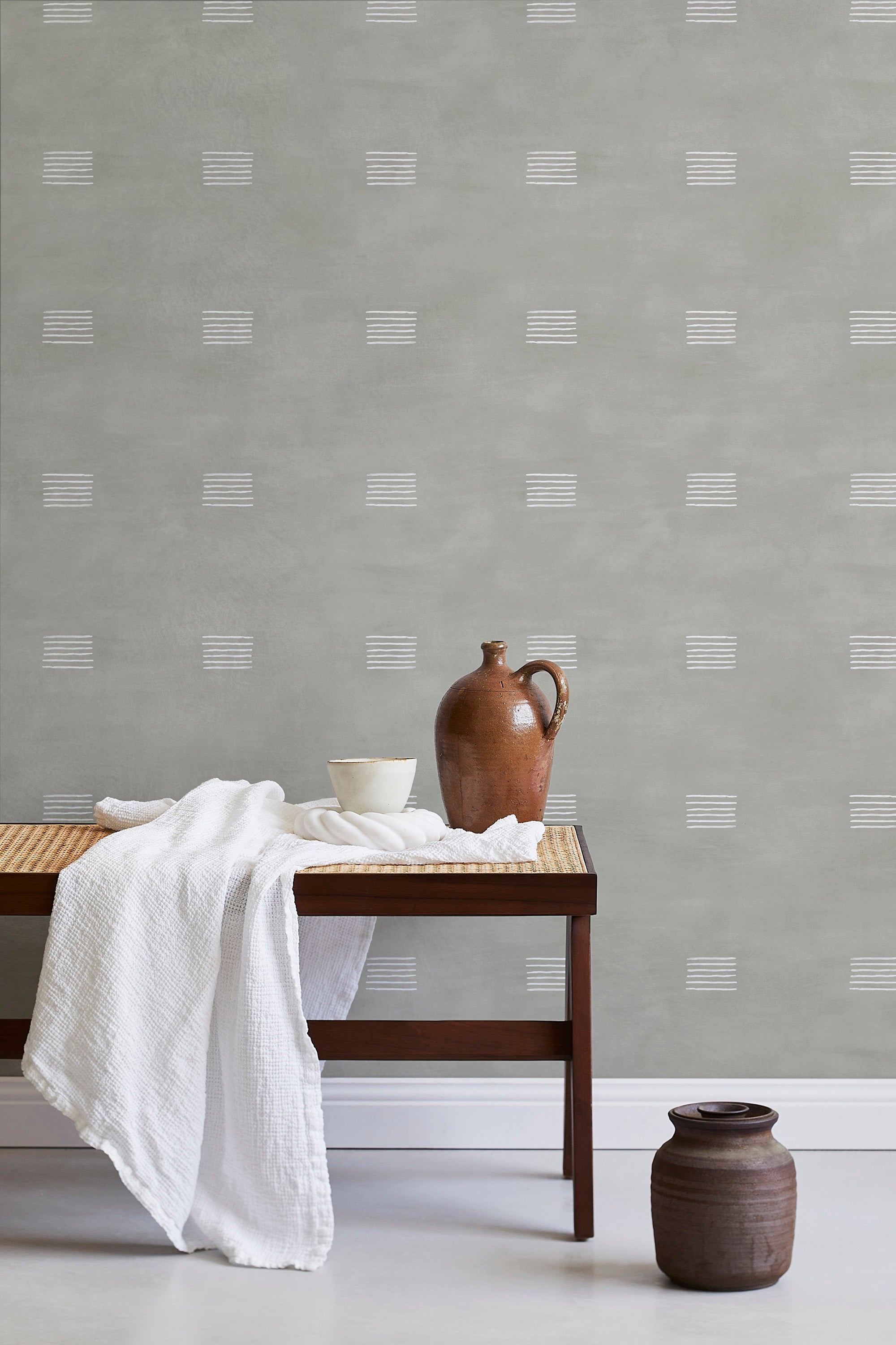 A bench with a towel and clay vases stands in front of a wall papered in a gridded dash print in white and sage.