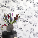 An end table with flowers stands in front of a wall papered in a playful illustrated city print in gray, black and white.