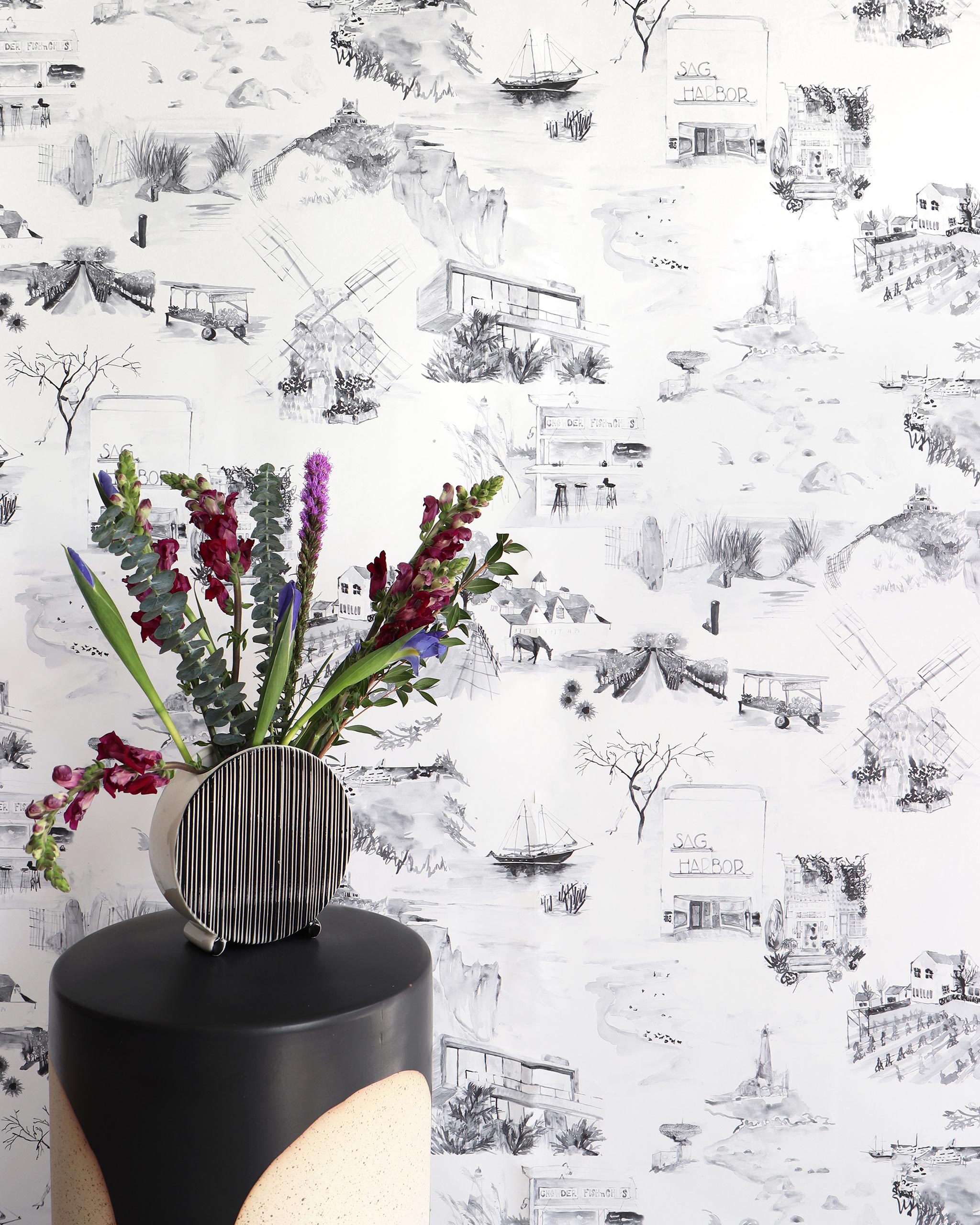 An end table with flowers stands in front of a wall papered in a playful illustrated city print in gray, black and white.