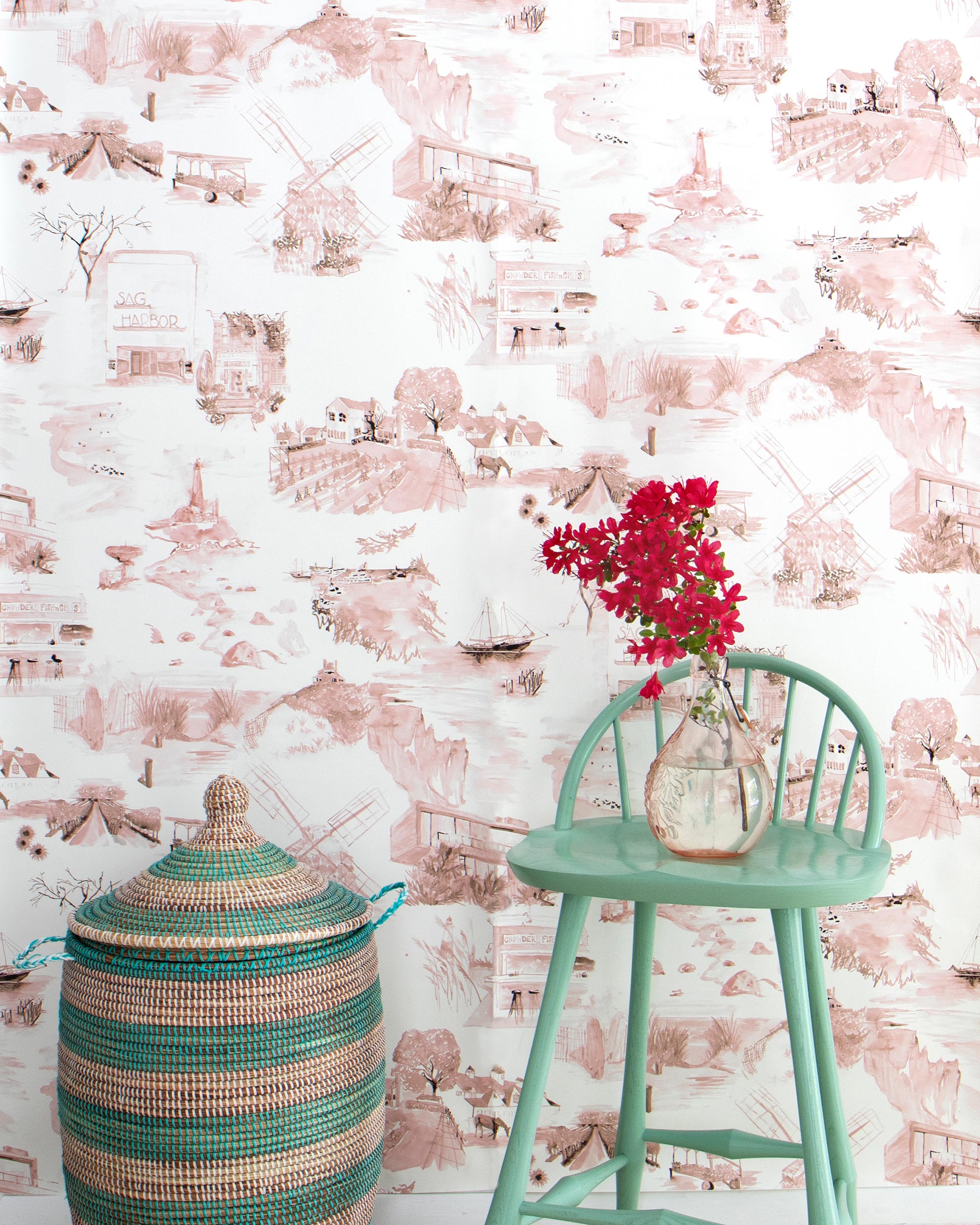 A chair, flowers and a basket stand in front of a wall papered in a playful illustrated city print in pink, brown and white.