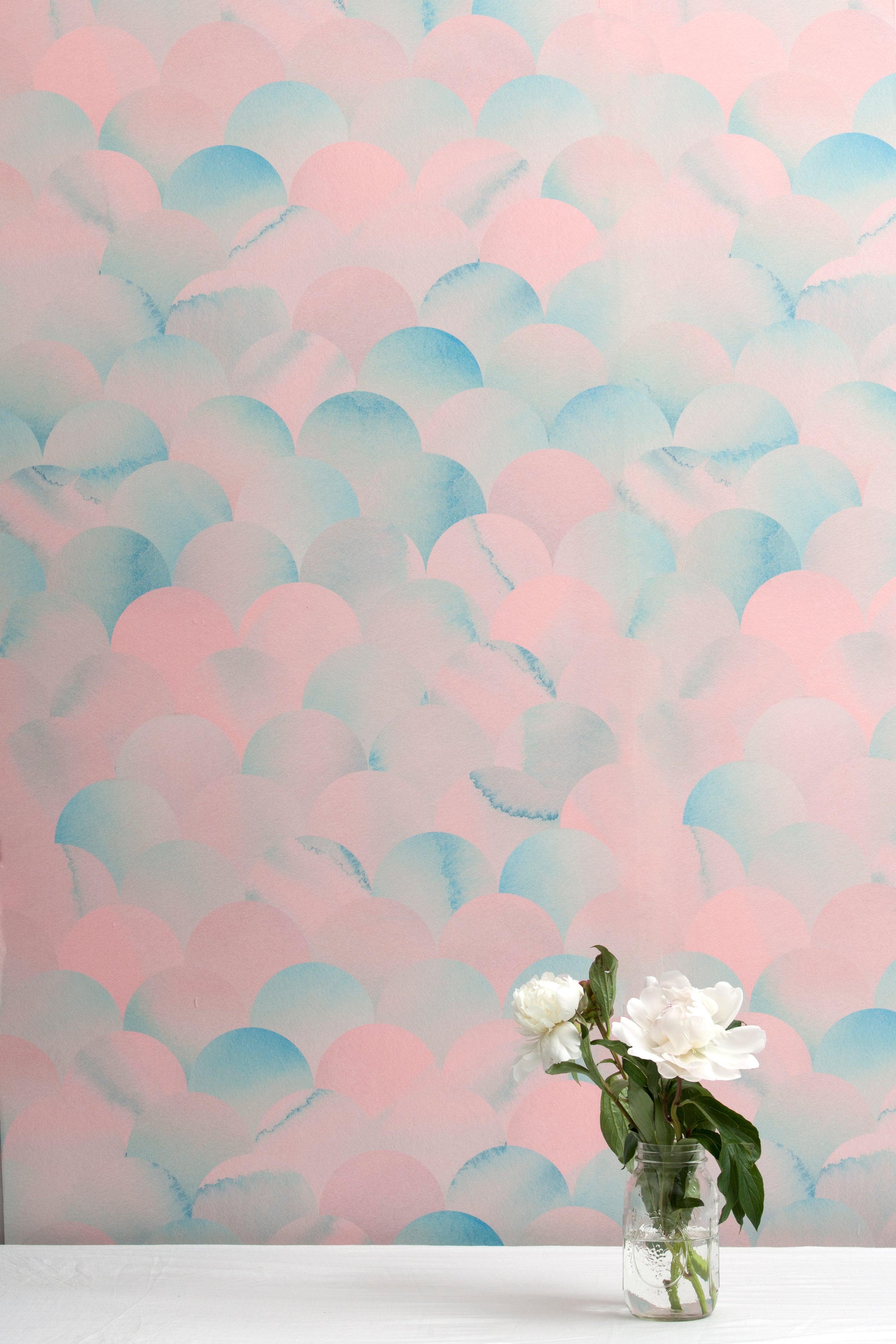 A vase of flowers stands in front of a wall papered in a repeating scalloped print in shades of blue and pink.