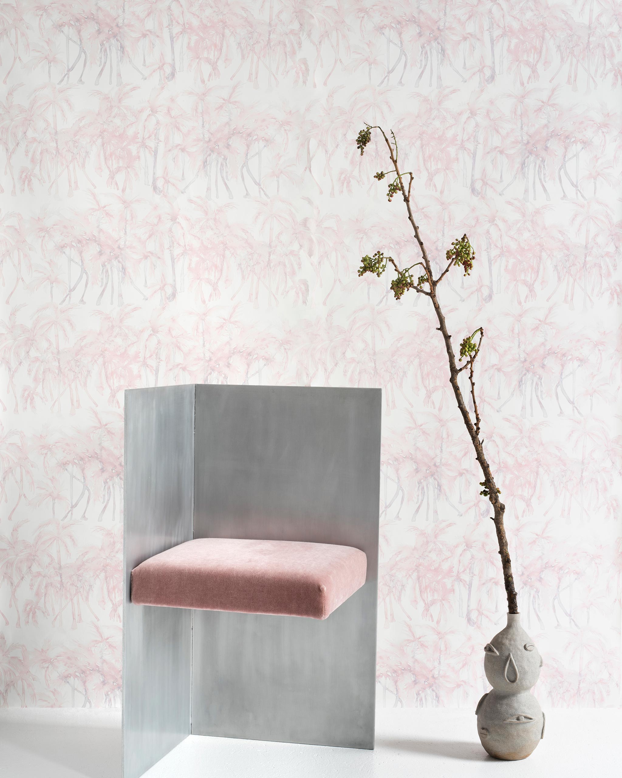 A modernist chair and plant stand in front of a wall papered in a painterly palm tree stripe print in pink, gray and white.