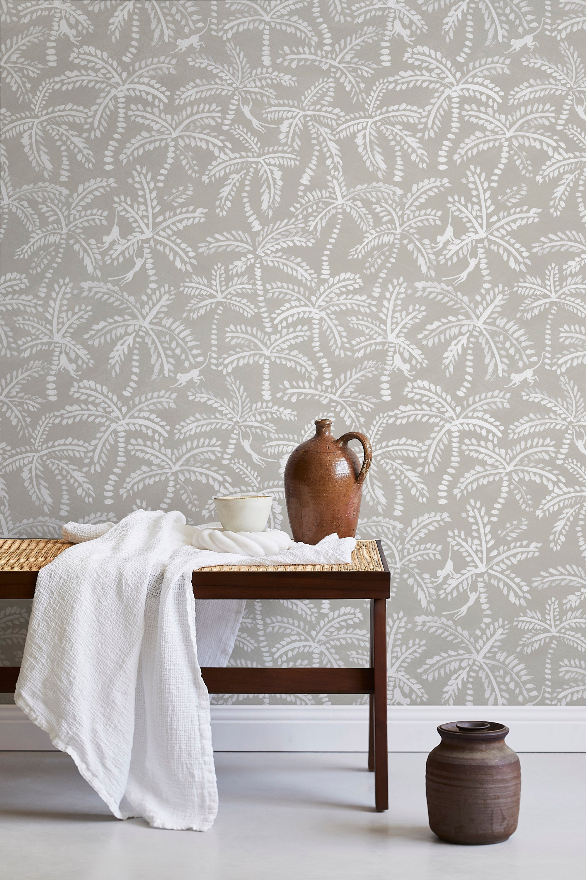 A bench with a towel and clay vases stands in front of a wall papered in a palm tree and monkey print in white and gray.