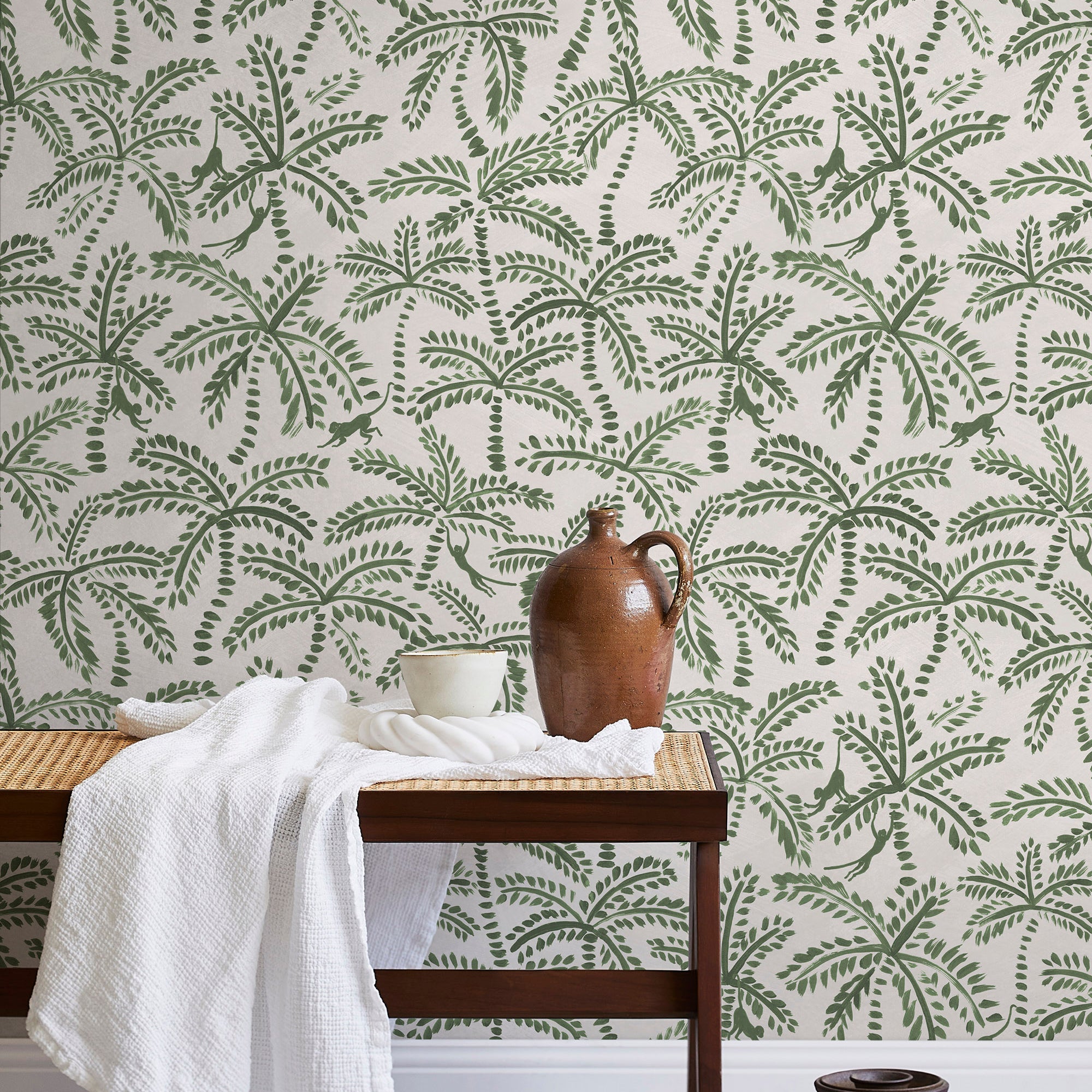 A bench with a towel and clay vases stands in front of a wall papered in a palm tree and monkey print in green and cream.