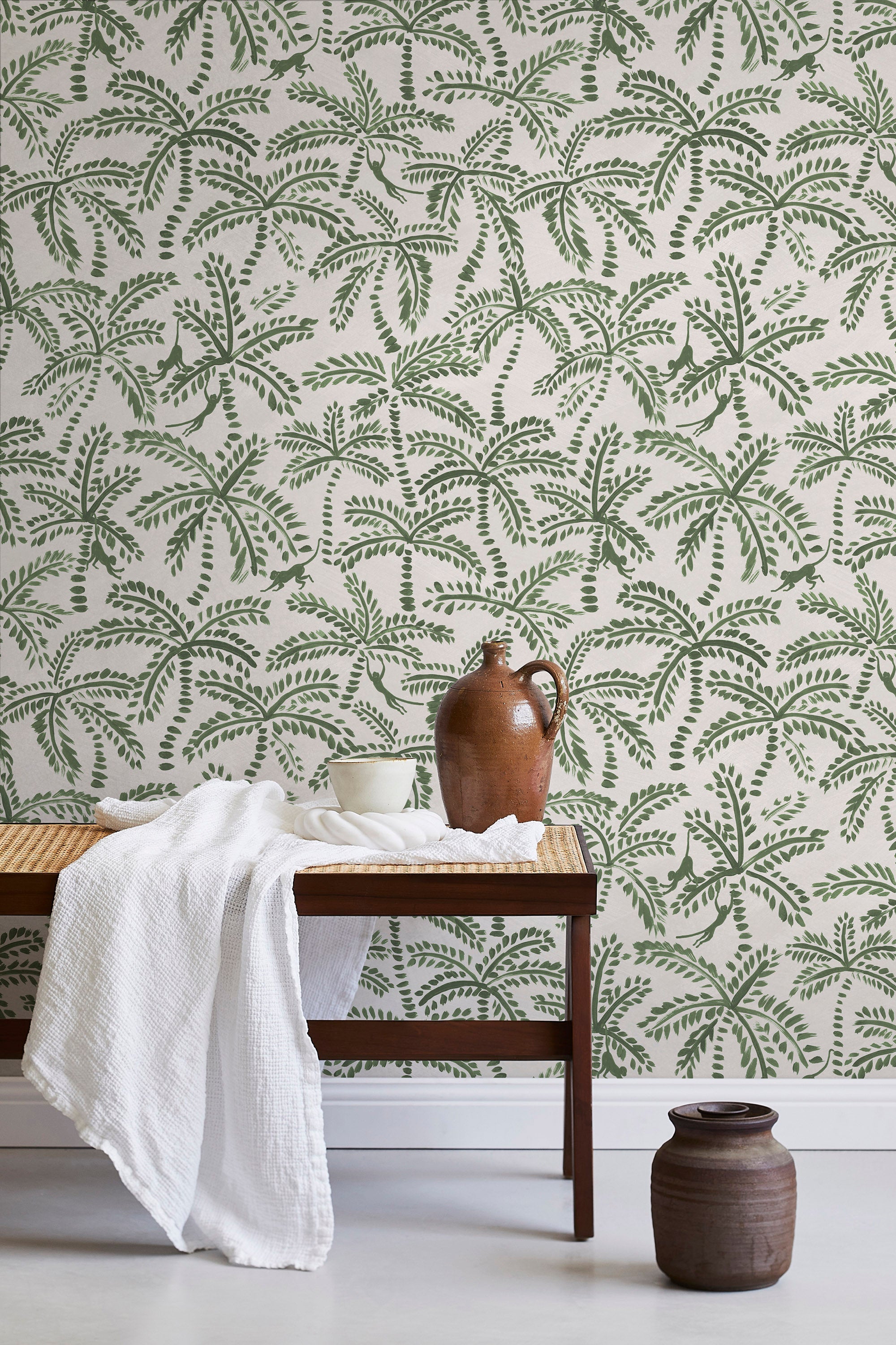 A bench with a towel and clay vases stands in front of a wall papered in a palm tree and monkey print in green and cream.