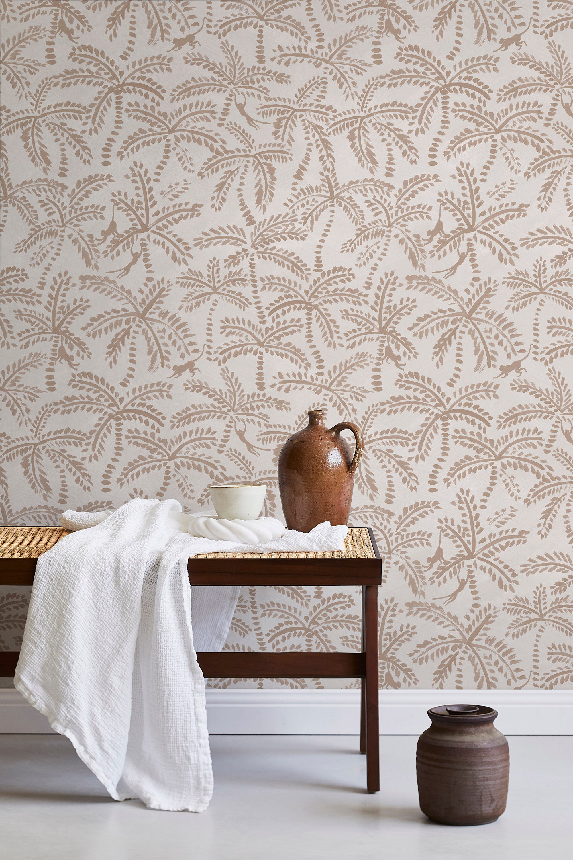 A bench with a towel and clay vases stands in front of a wall papered in a palm tree and monkey print in tan and cream.