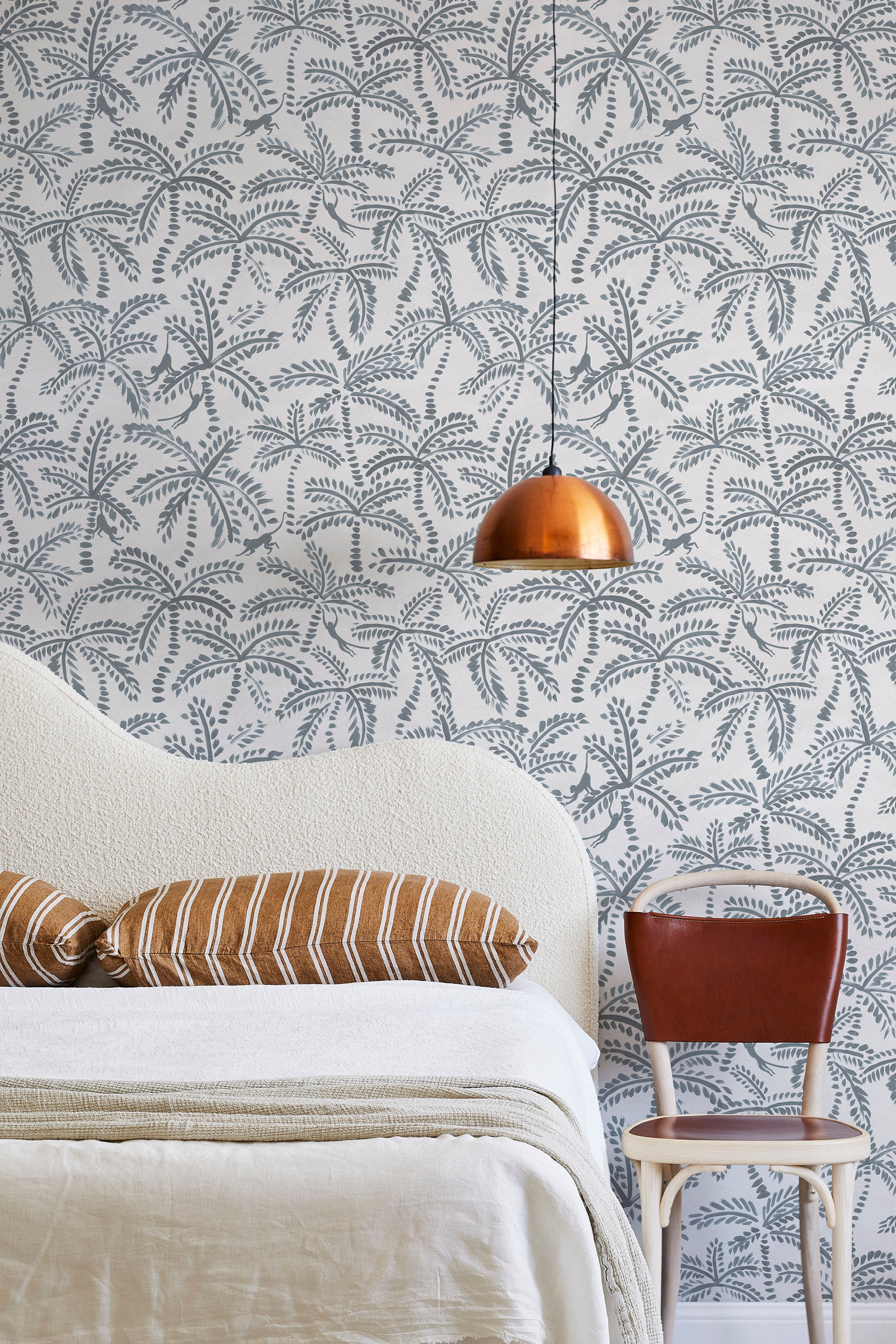 A modernist bed, hanging lamp and chair stand in front of a wall papered in a palm tree and monkey print in gray and white.