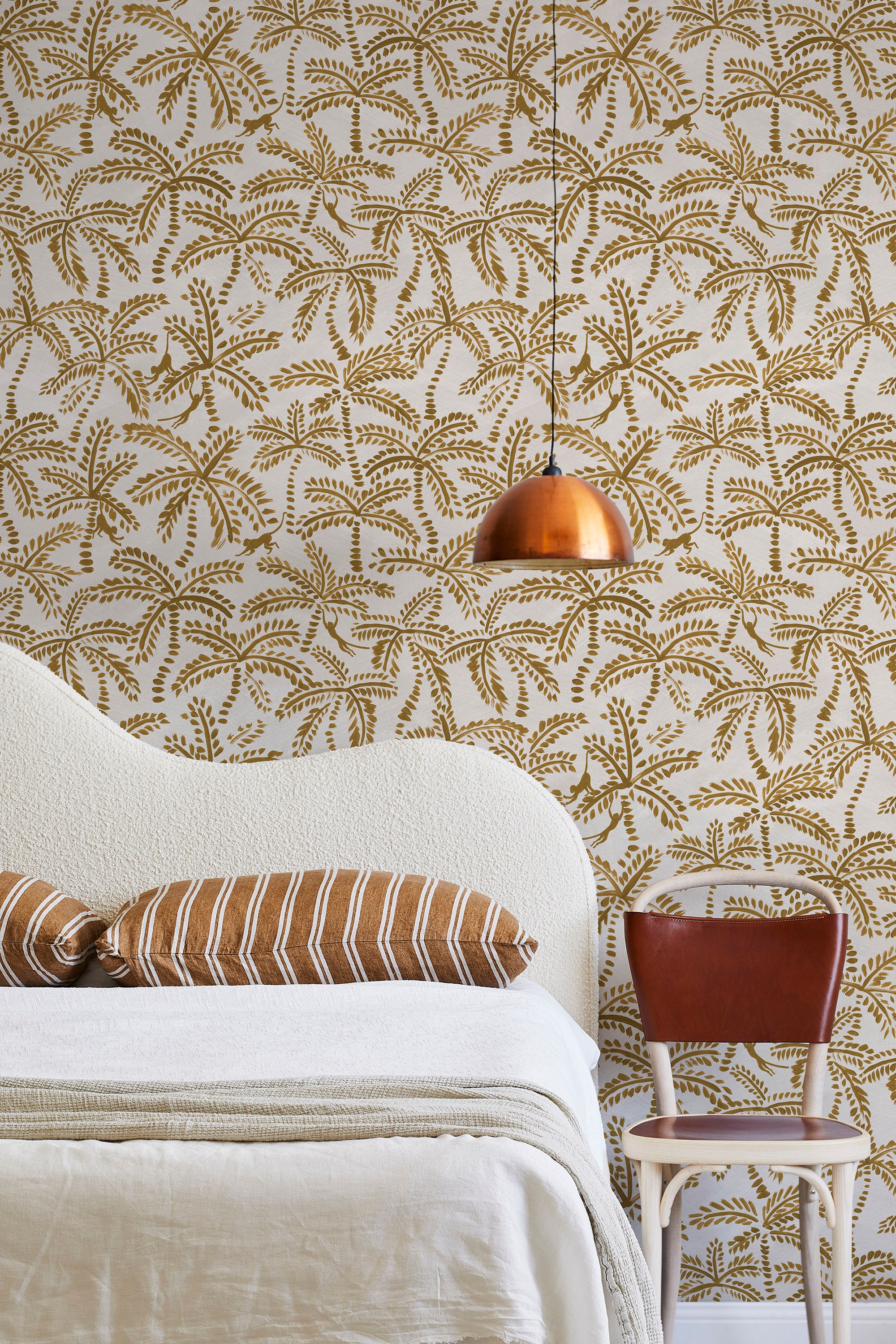 A modernist bed, hanging lamp and chair stand in front of a wall papered in a palm tree and monkey print in gold and cream.