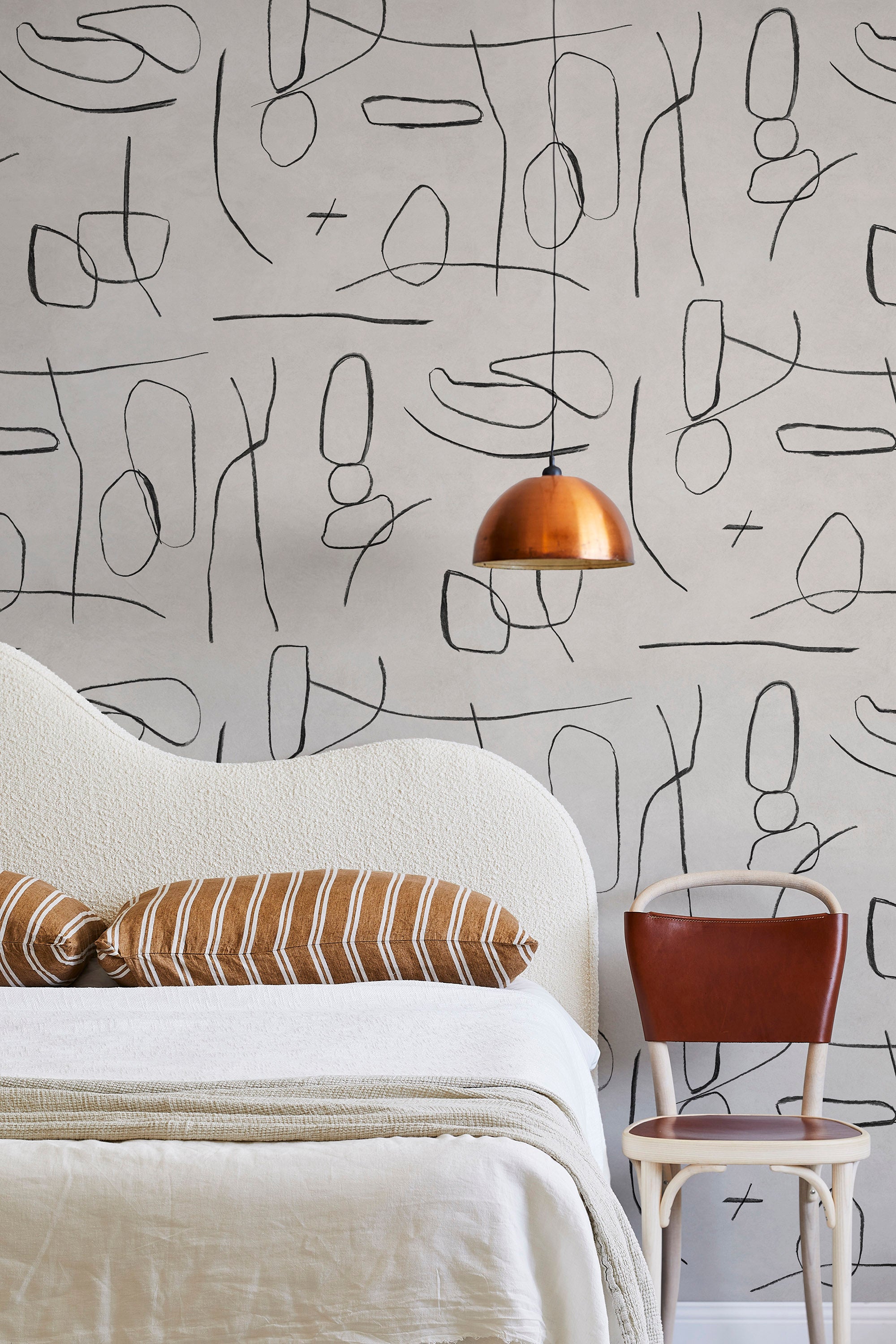 A modernist bed, hanging lamp and chair stand in front of a wall papered in an abstract shape pattern in black and tan.