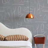 A modernist bed, hanging lamp and chair stand in front of a wall papered in an abstract shape pattern in white and gray.