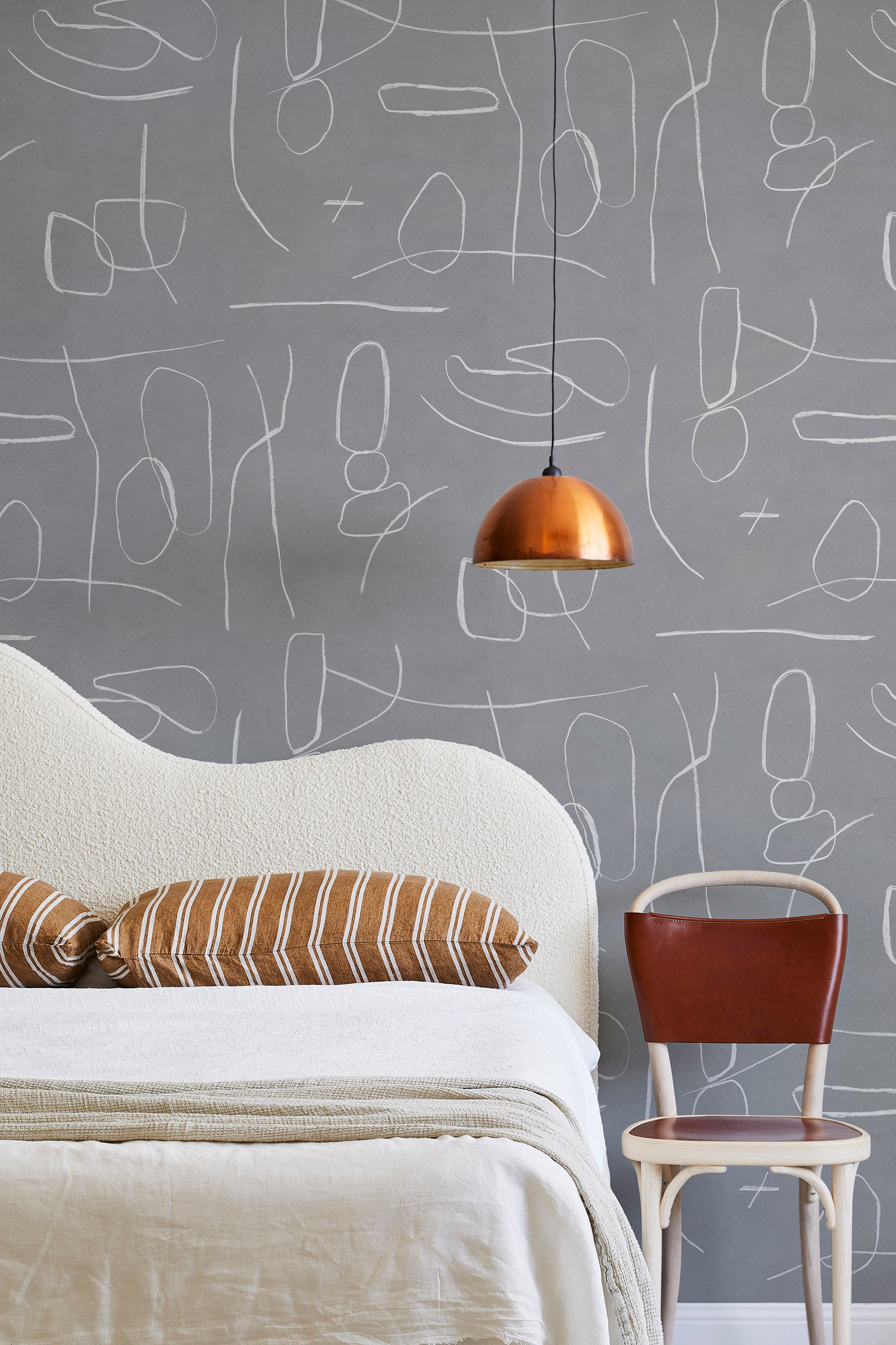 A modernist bed, hanging lamp and chair stand in front of a wall papered in an abstract shape pattern in white and gray.