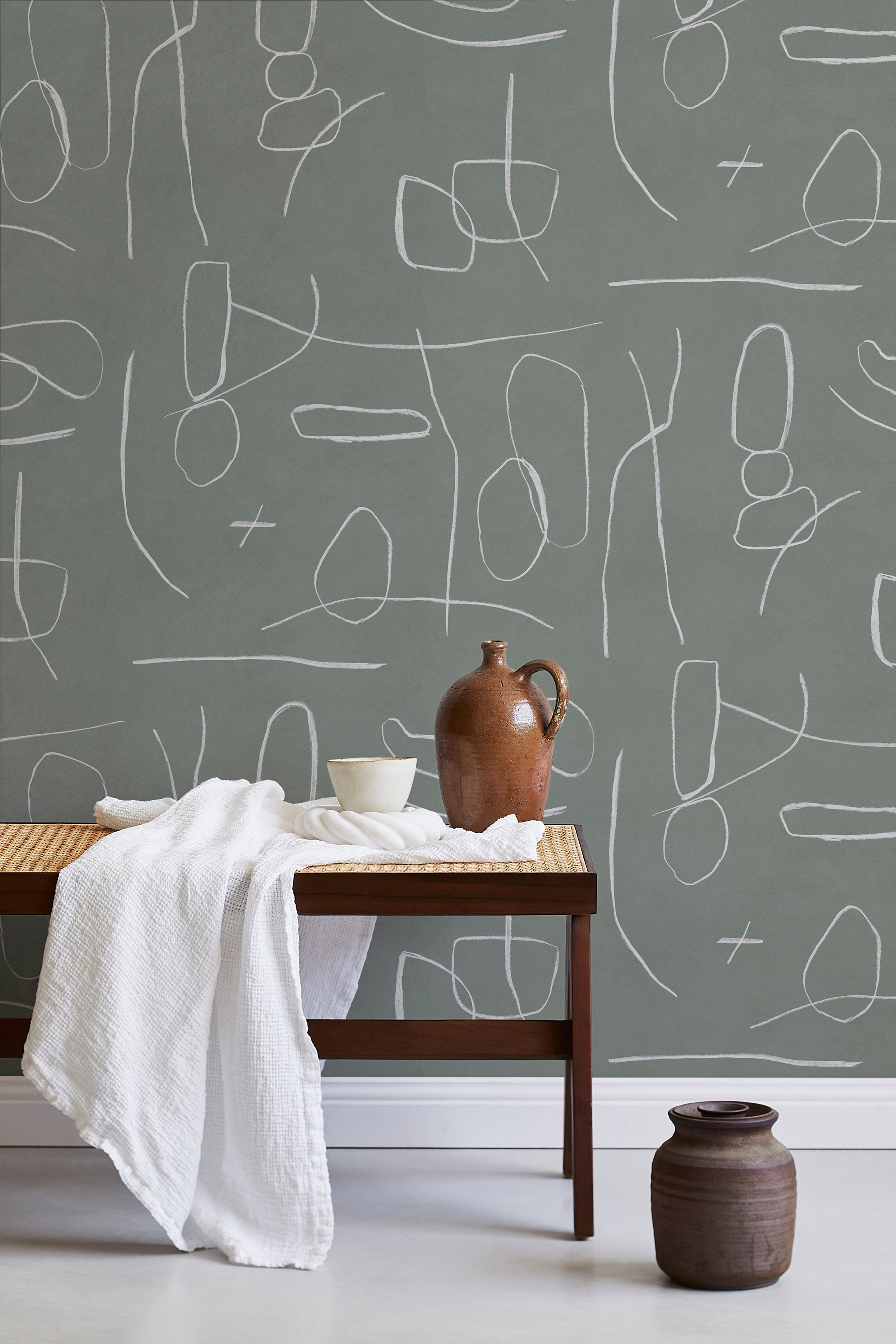 A bench with a towel and clay vases stands in front of a wall papered in an abstract shape pattern in white and sage.