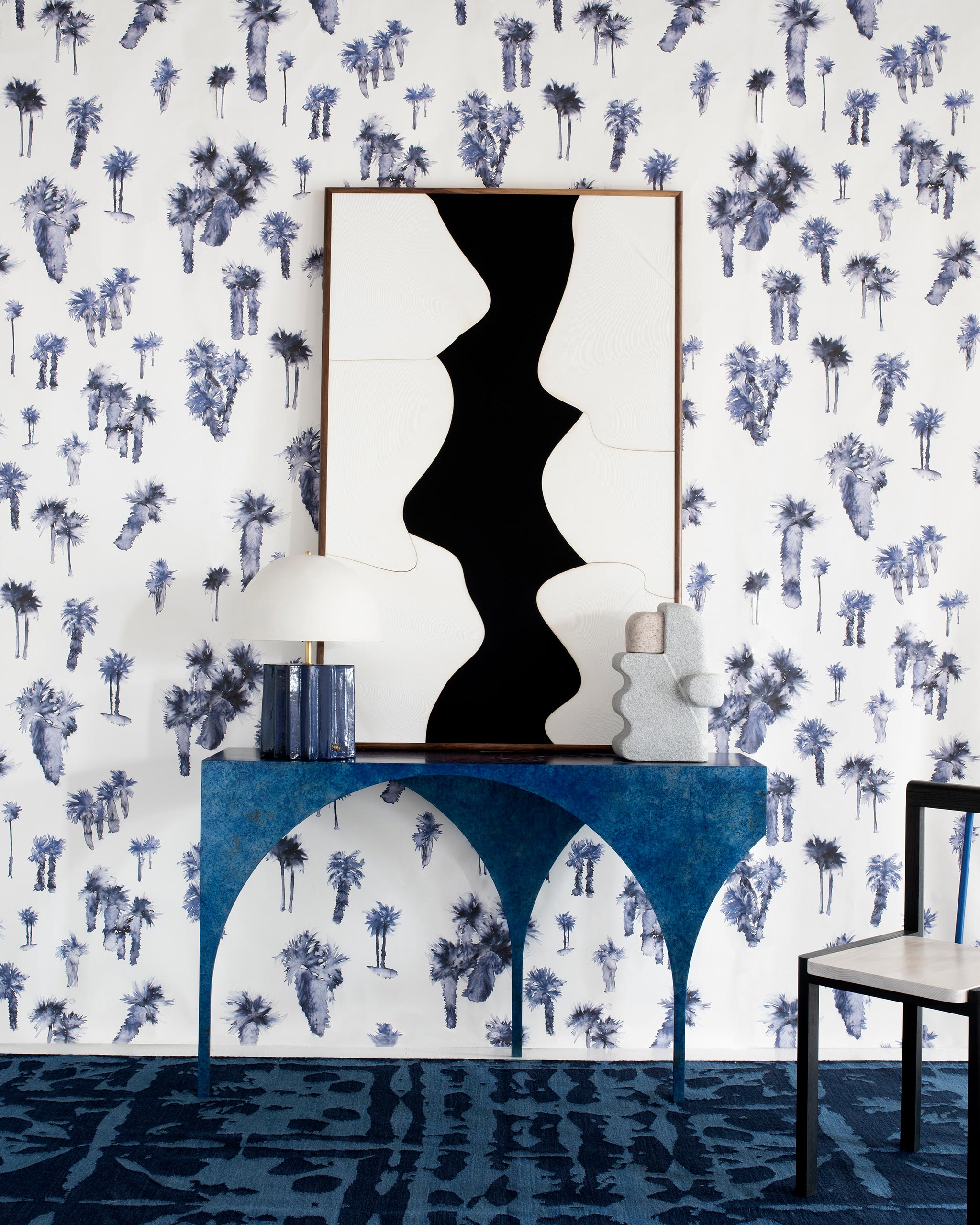 A modernist end table with art stands in front of a wall papered in a playful palm tree print in navy, gray and white.