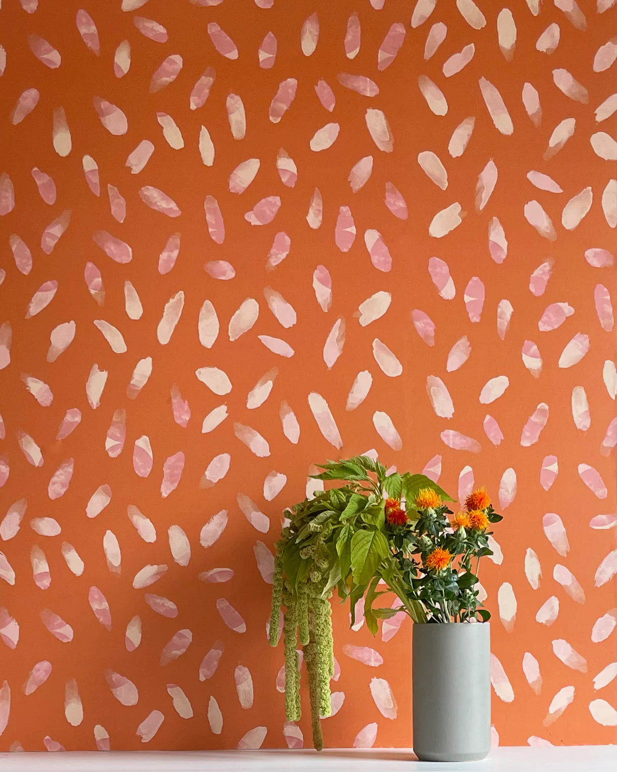 A vase of flowers stands in front of a wall covered in a playful paint blotch print in pink and cream on an orange field.