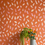 A vase of flowers stands in front of a wall covered in a playful paint blotch print in pink and cream on an orange field.