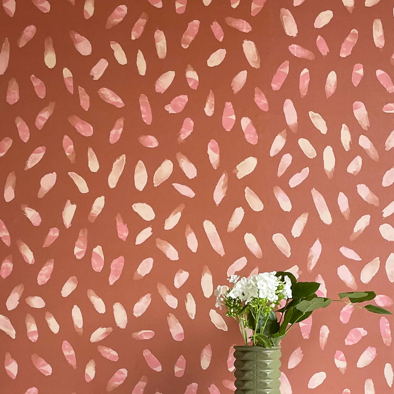 A vase of flowers stands in front of a wall covered in a playful paint blotch print in pink and cream on a brown field.