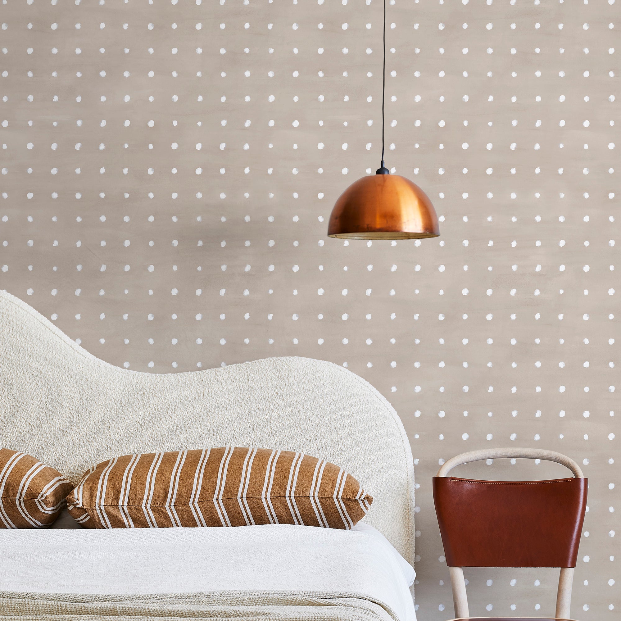 A modernist bed, hanging lamp and chair stand in front of a wall papered in a dotted grid pattern in white and tan.