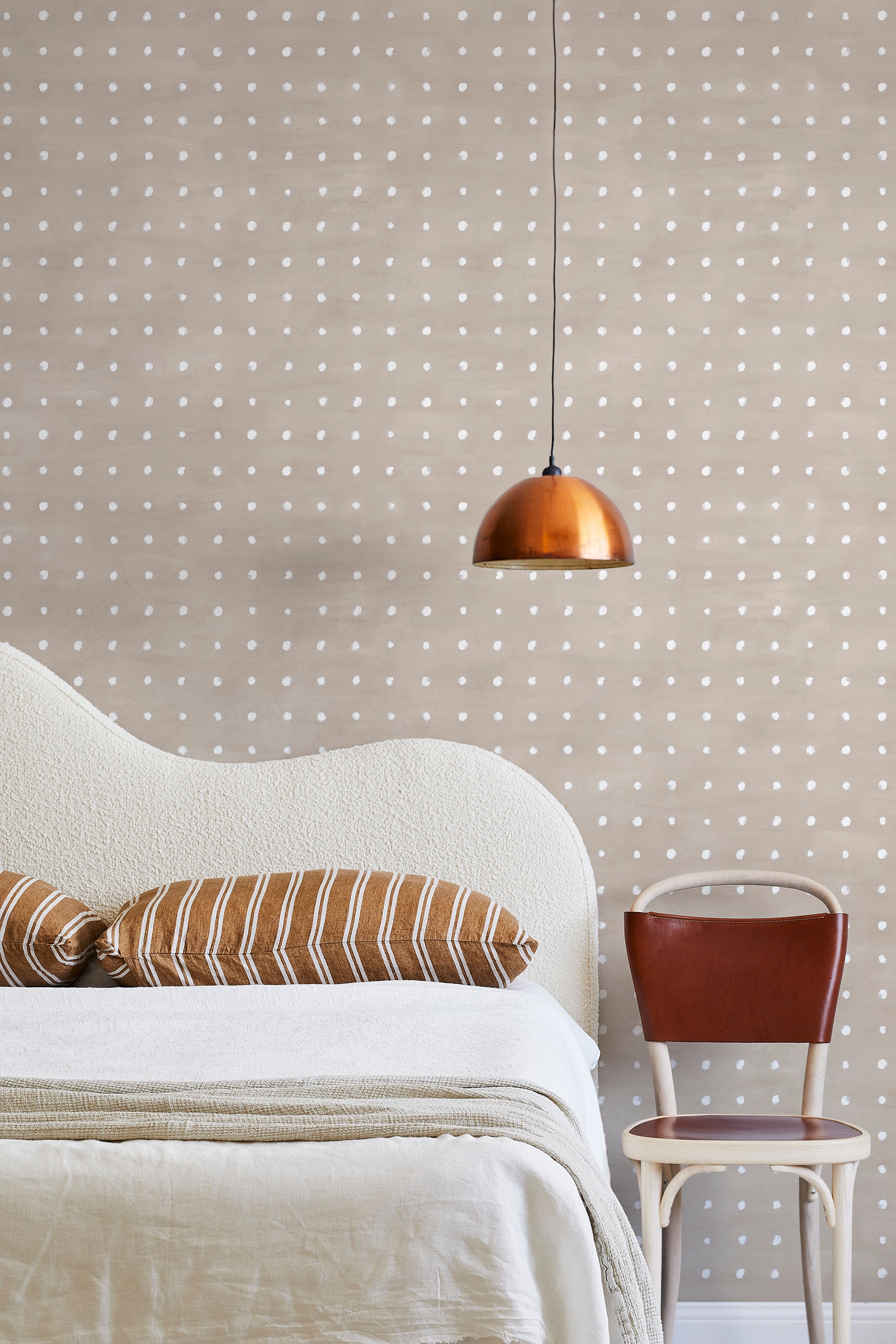 A modernist bed, hanging lamp and chair stand in front of a wall papered in a dotted grid pattern in white and tan.