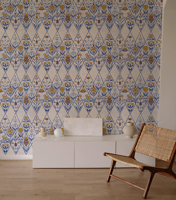 An armchair and credenza stand in front of a wall papered in a floral and diamond lattice print in blue, gold and white.