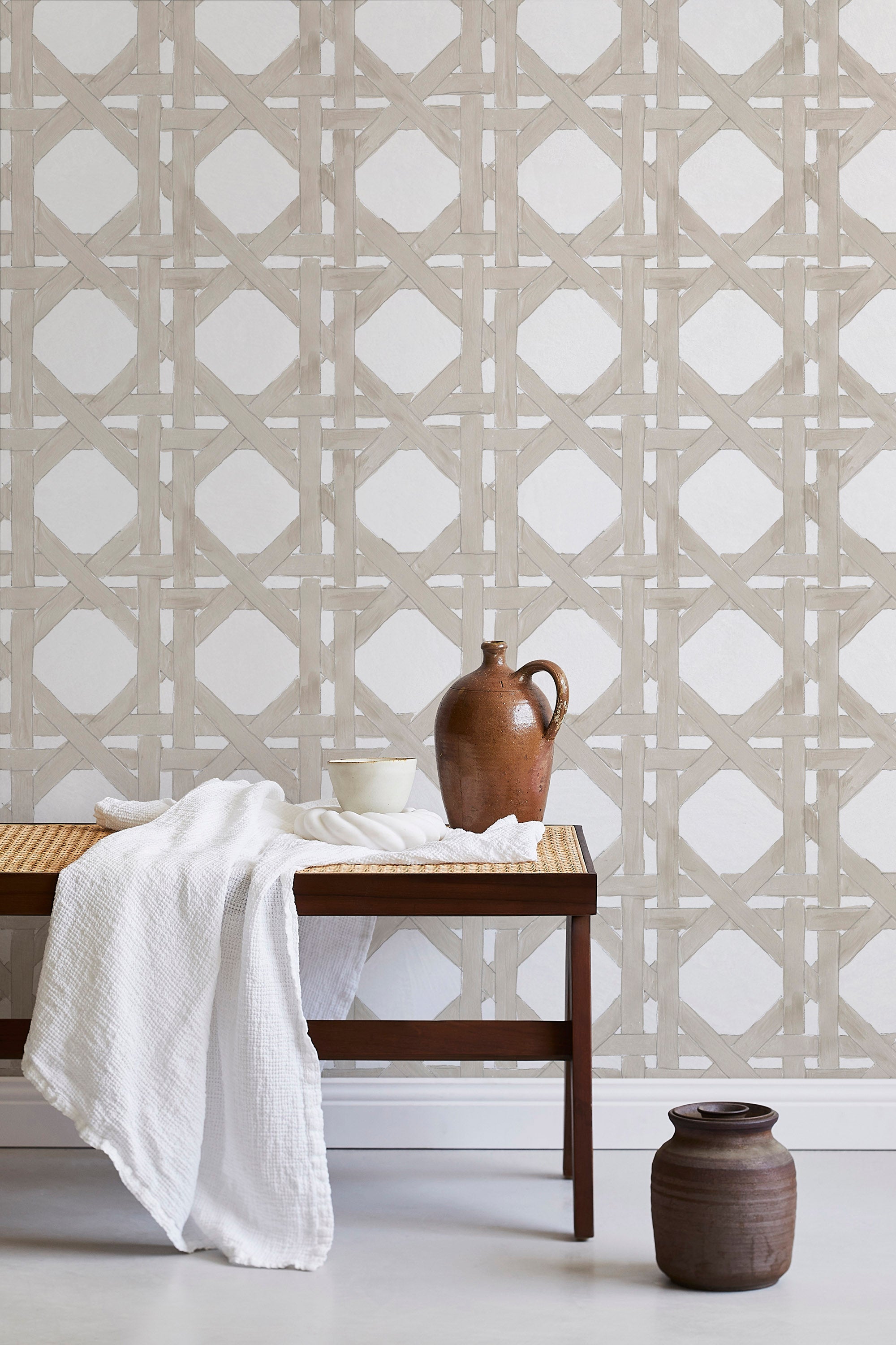 A bench with a towel and clay vases stands in front of a wall papered in a geometric lattice print in tan and white.