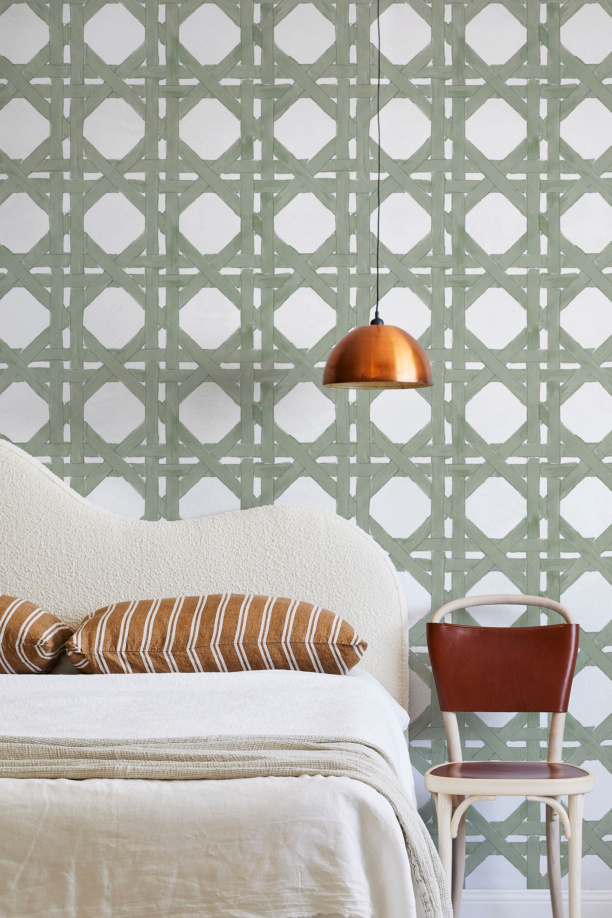 A modernist bed, hanging lamp and chair stand in front of a wall papered in a geometric lattice print in sage and white.